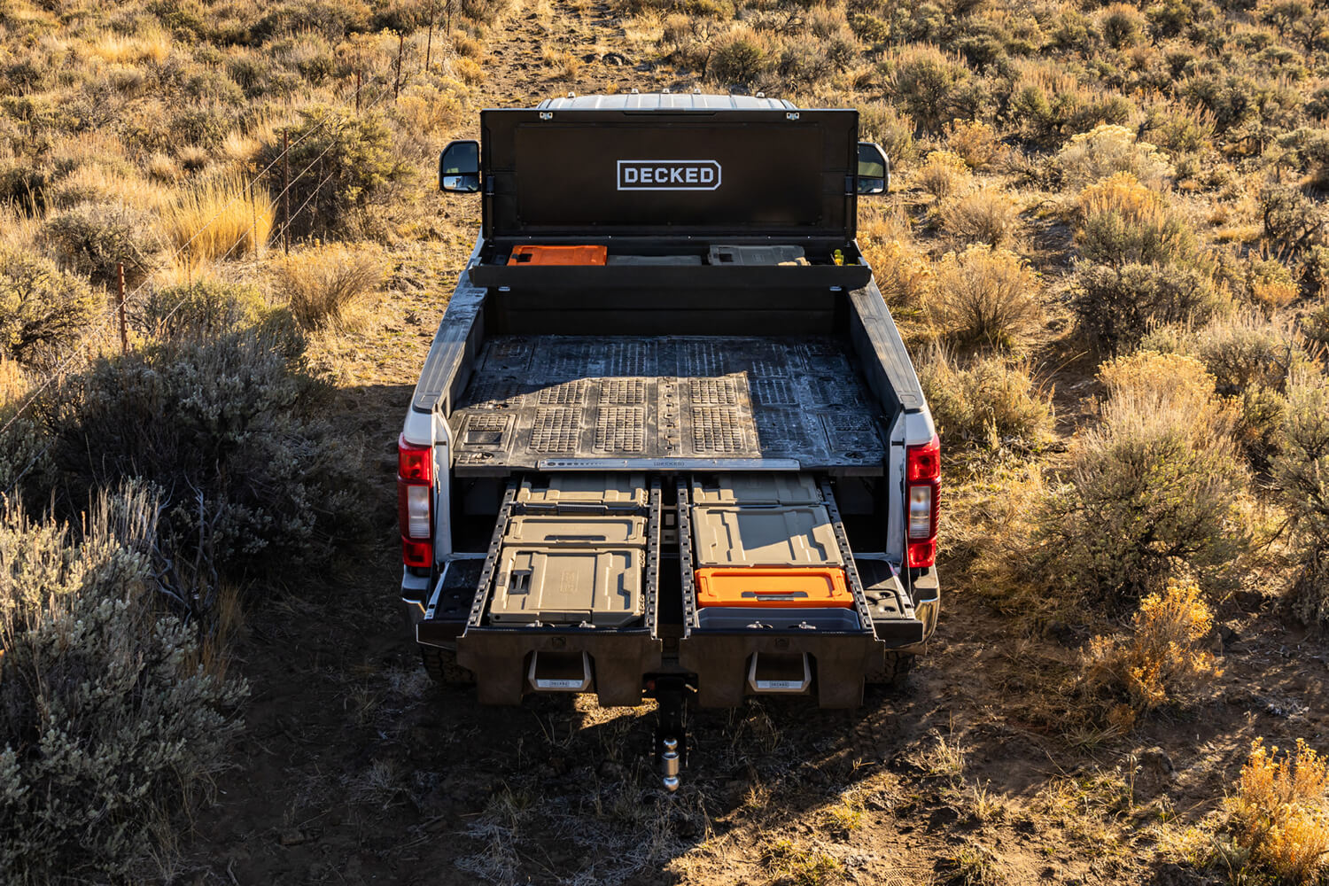 Open black DECKED toolbox and drawer system showing D-CO case storage in the bed of a Ford truck parked in a field.