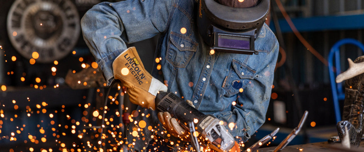 A welder in a blue denim work jacket and protective helmet sparks fly as they use a Lincoln Electric grinder, with bright orange sparks glowing in the foreground.
