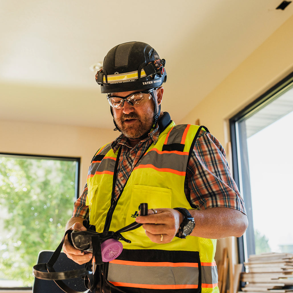 Construction worker wearing a hard hat and high-visibility vest prepares a respirator inside a building.