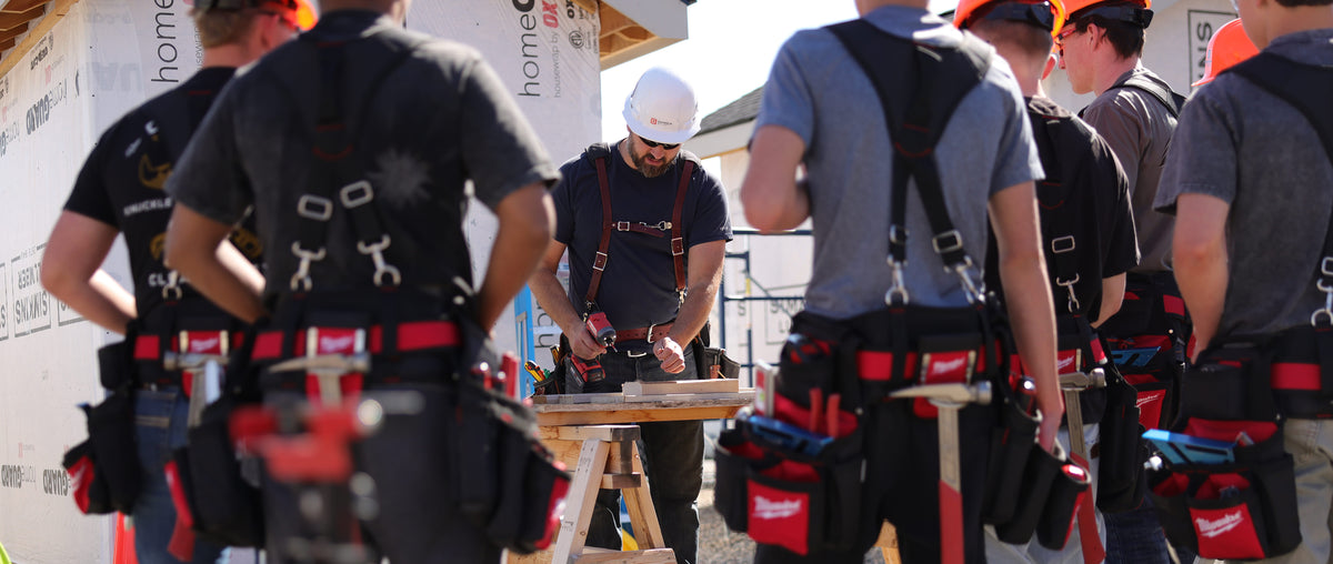 A carpenter works on lumber at an outdoor jobsite while multiple members look on.