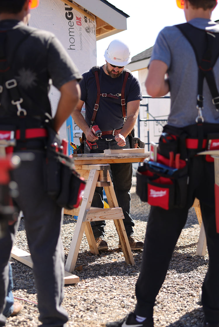 A carpenter works on lumber at an outdoor jobsite while multiple members look on.