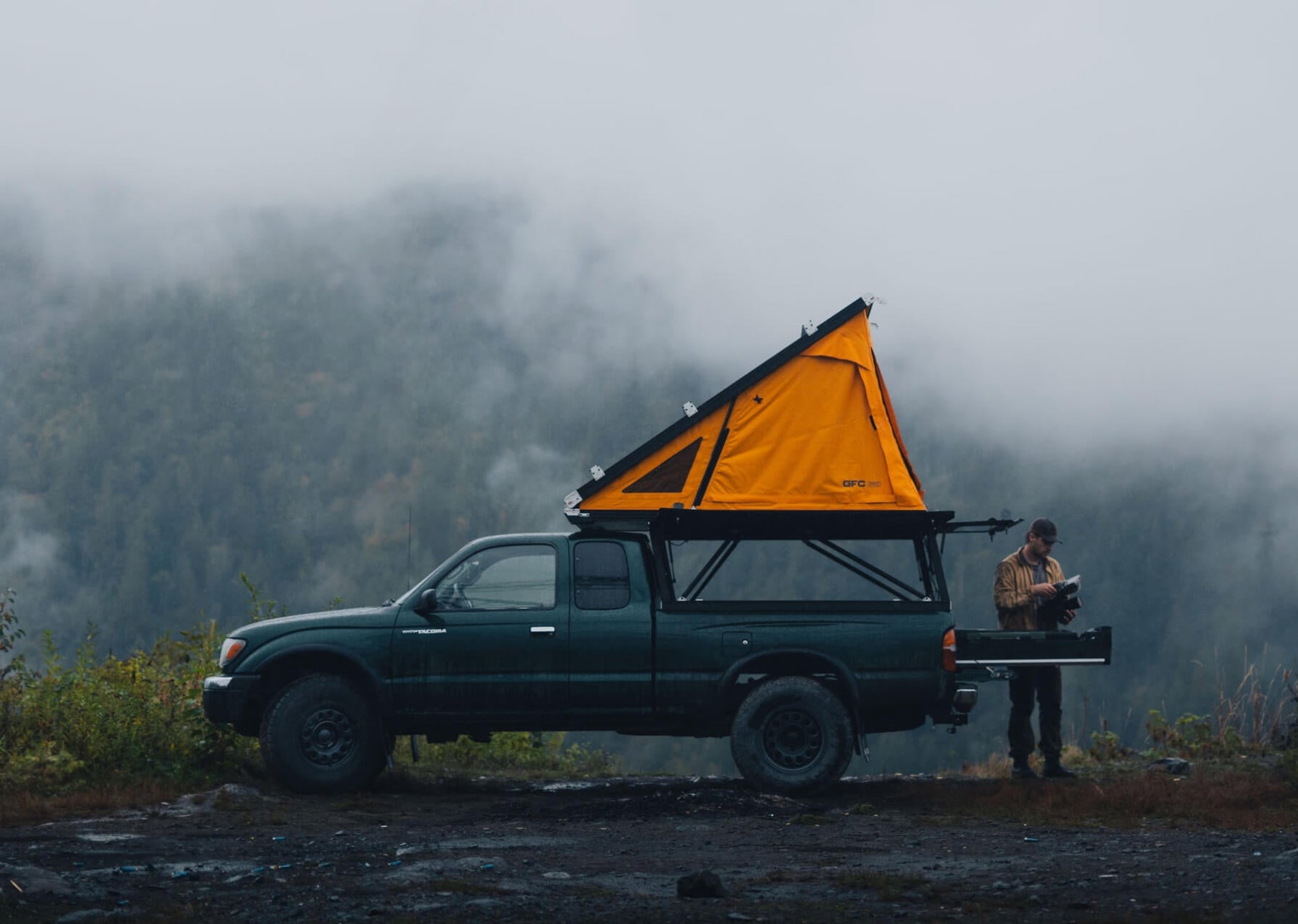 Person standing next to a truck with an orange rooftop tent in a foggy forest.