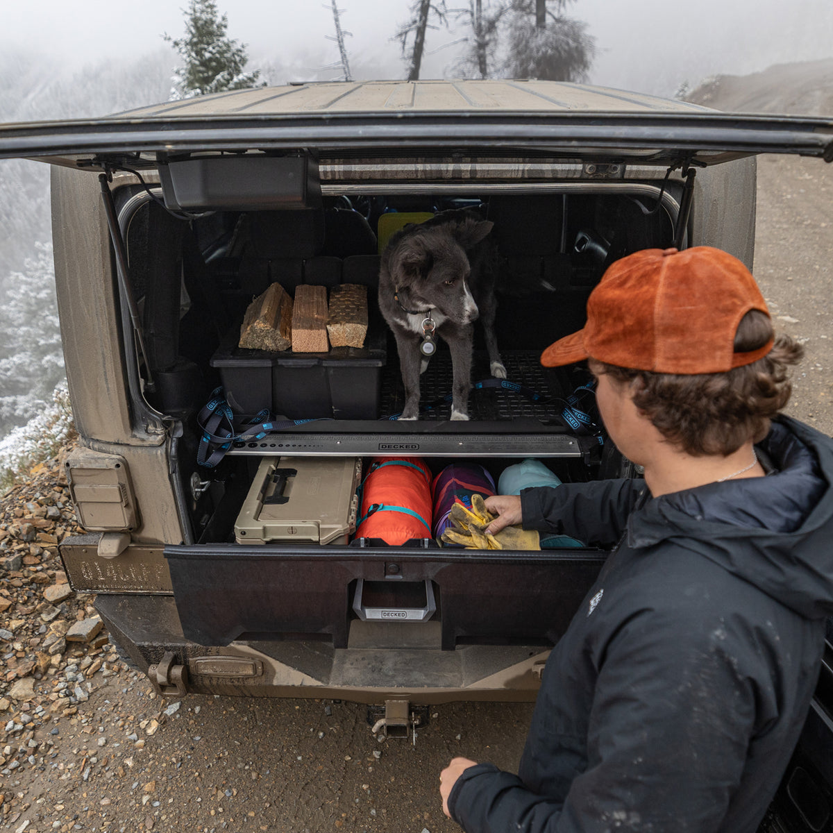 Man standing beside an open DECKED drawer filled with gear while a dog sits in the back of a Jeep Wrangler.