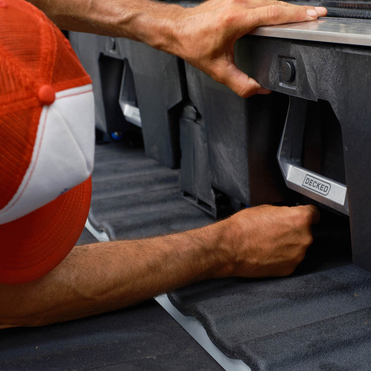 A close-up of a person installing a black decked drawer system into a truck bed. The person is wearing a red and white trucker hat.