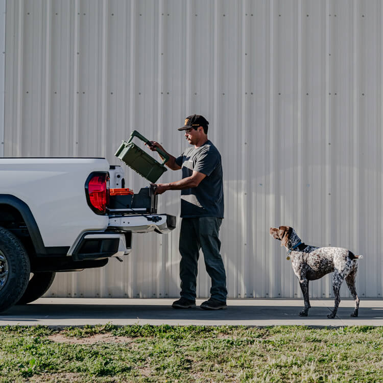 Man loading a protective case into a truck bed's DECKED Drawer System with a dog standing nearby, against a gray building.