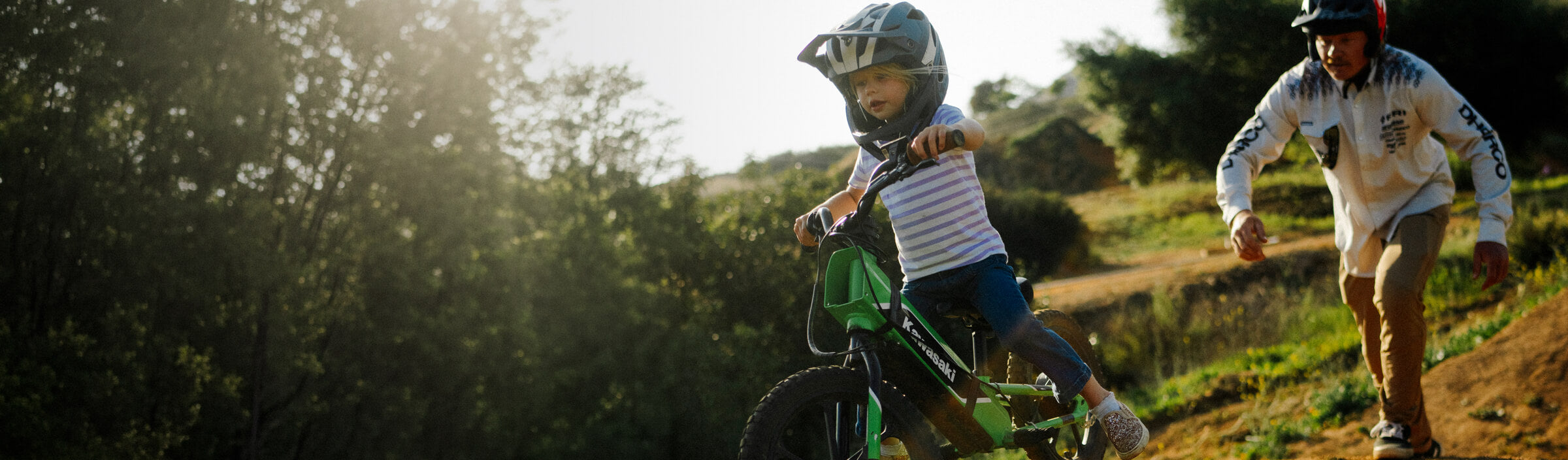 A little girl riding a green balance bike on a dirt single track with her dad chasing after her.