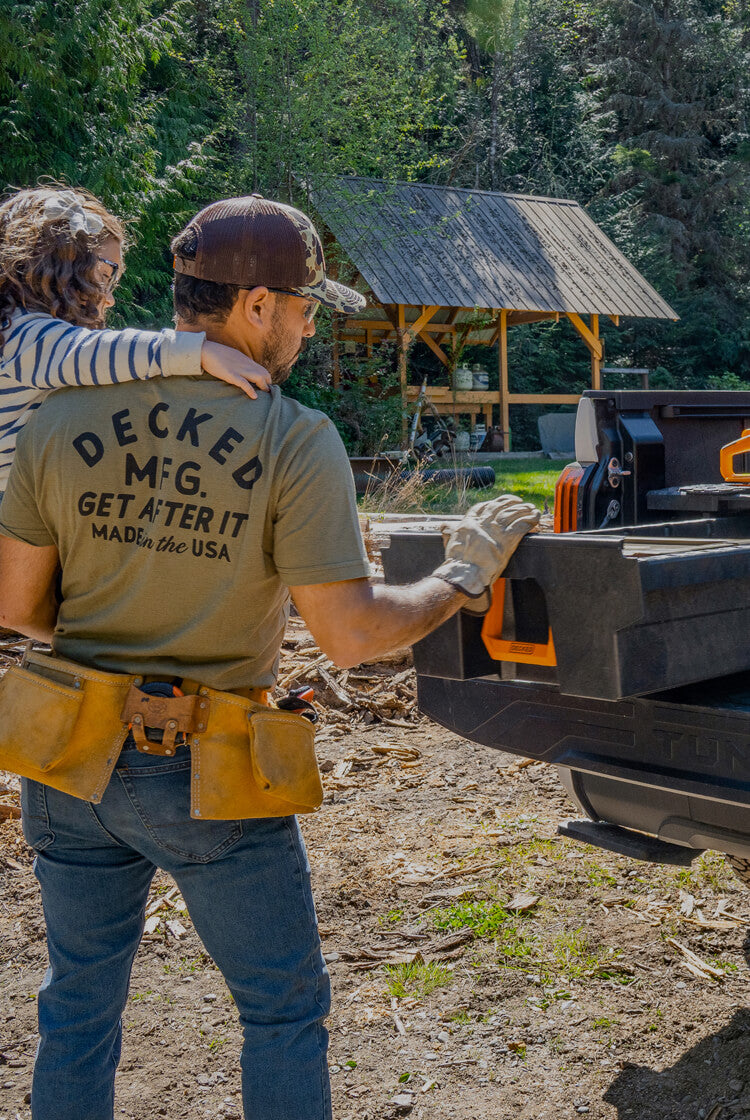 A dad and his daughter putting tools away into his DECKED Drawer System with orange Hop Up Kit handles in a wooded area with a truck bed filled with logs and equipment.
