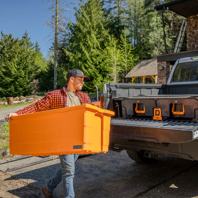 A man loading a large orange Payloader 133 Bin into the back of his truck bed that has a DECKED Drawer System inside.