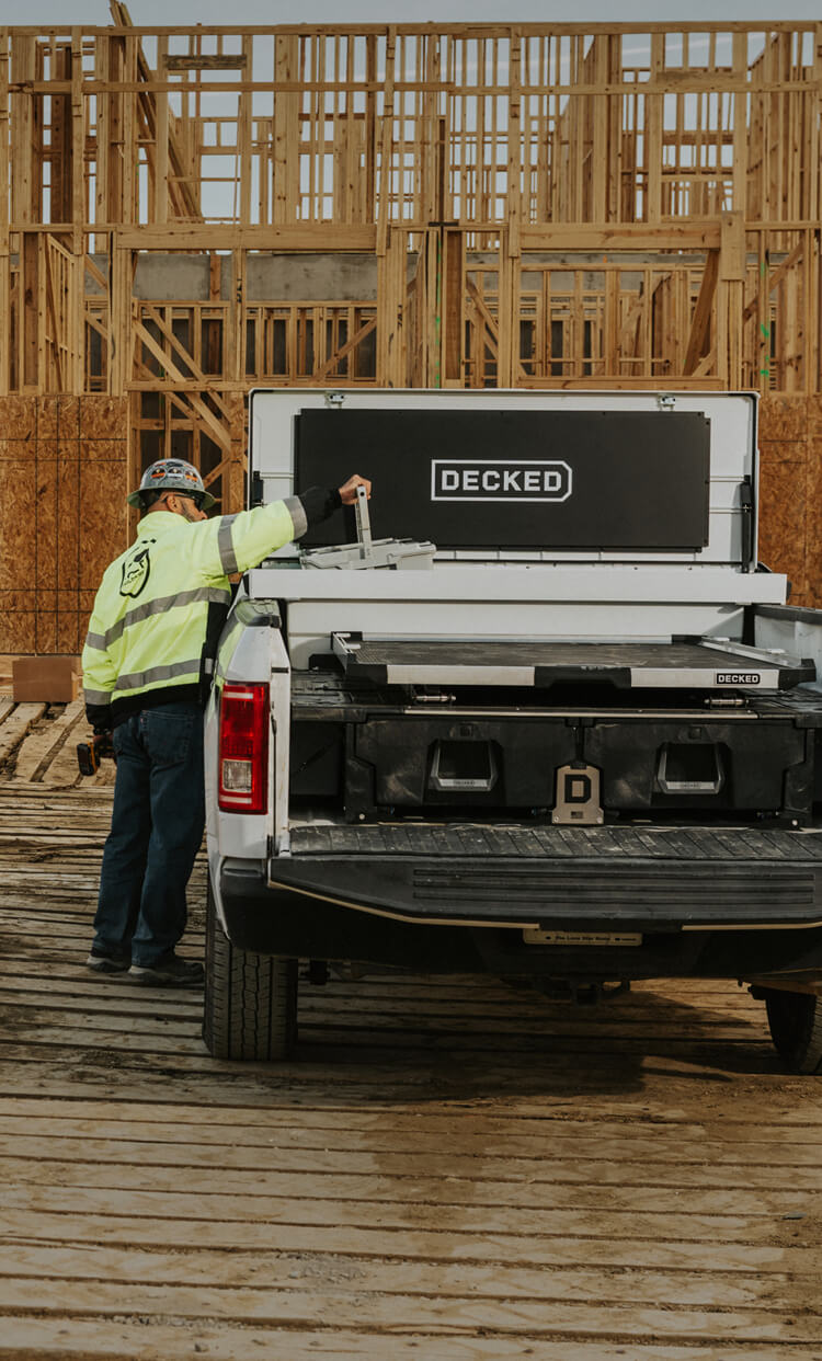 Person standing next to a truck with a 'DECKED' Tool Box, Drawer System, and CargoGlide at a construction site.