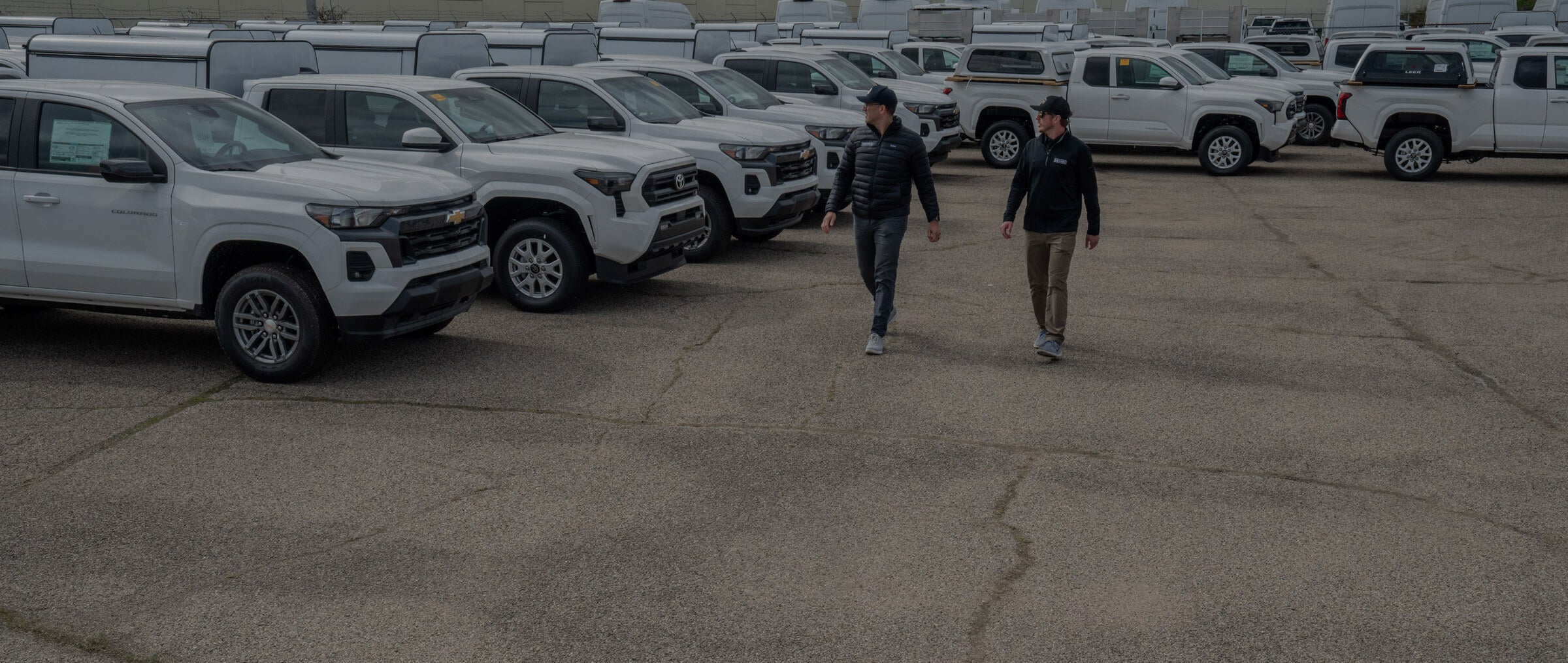 Two people walking between white pickup trucks in a row.
