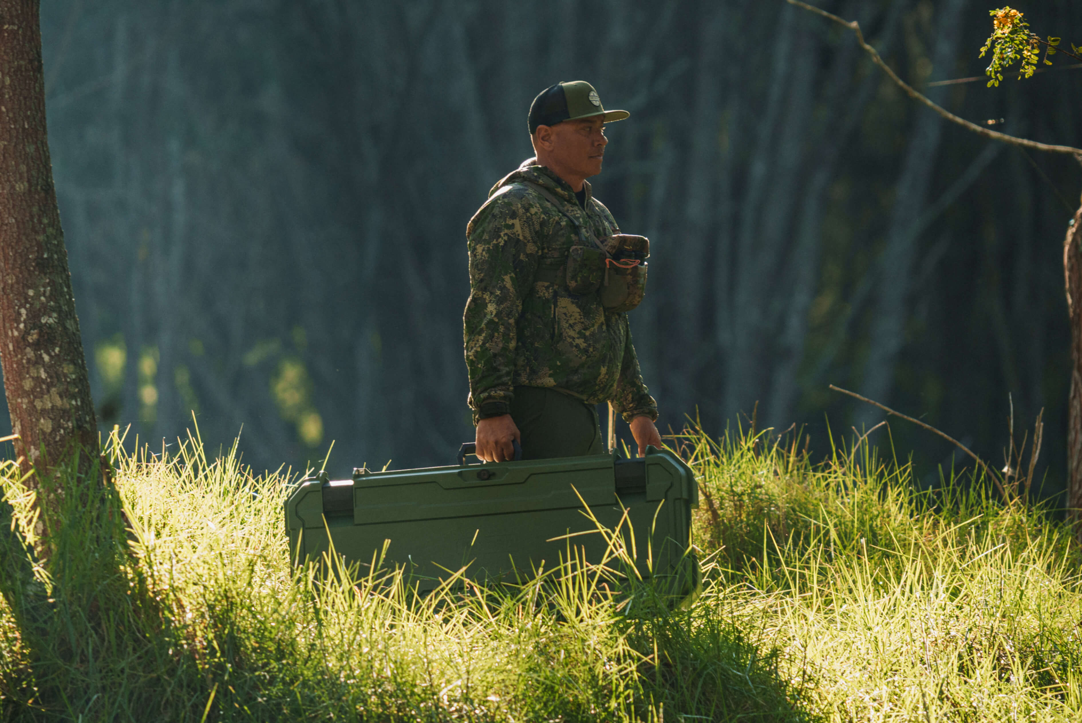 A man in camo carrying his Ranger Green Honcho with his bow inside through the woods.
