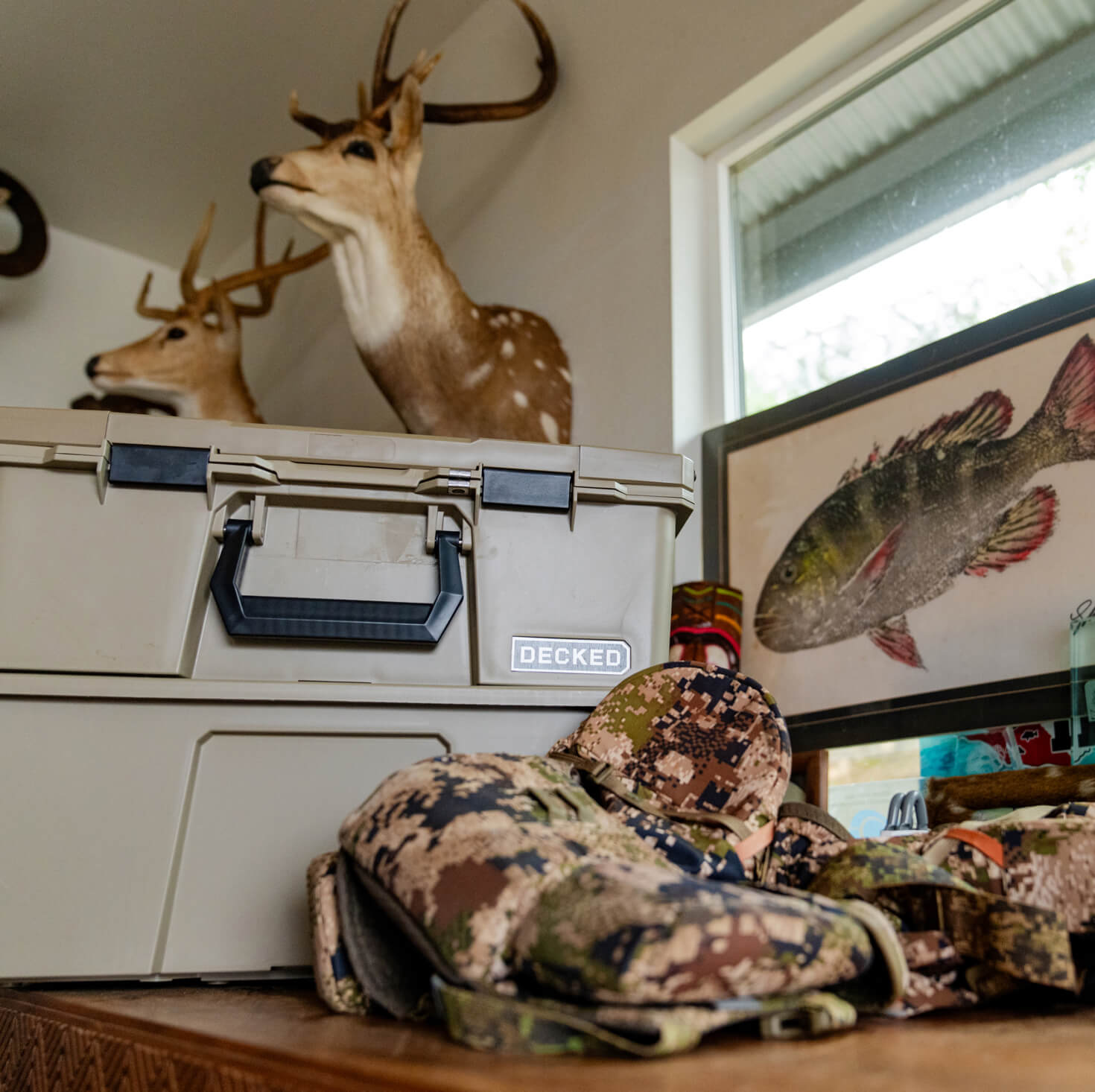 Camouflage jacket on a work bench next to two desert tan DECKED Cases and Bins with deer heads mounted on the wall.