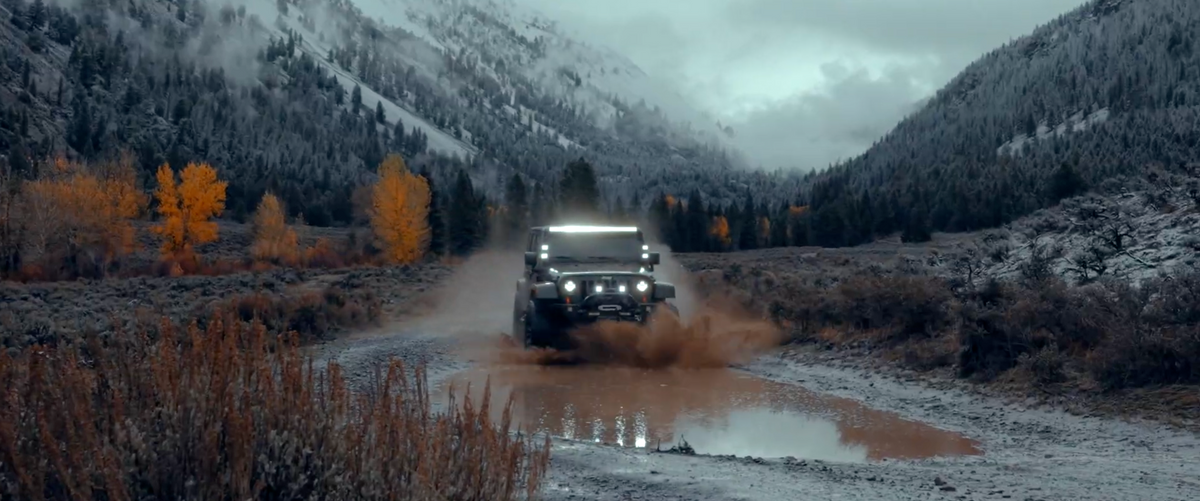 Jeep Wrangler driving through a muddy puddle on a road surrounded by snow-dusted mountains and fall foliage.