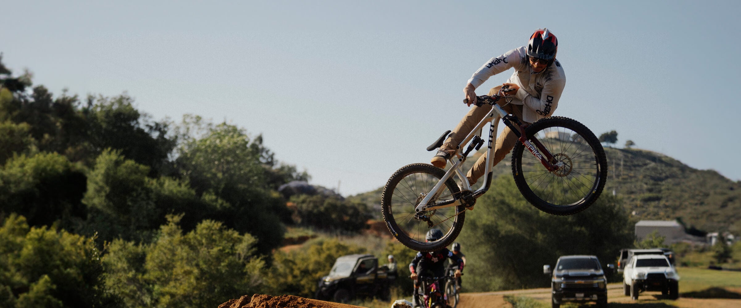 Kyle Strait on a bicycle performing a jump in an outdoor setting with trees and vehicles in the background.
