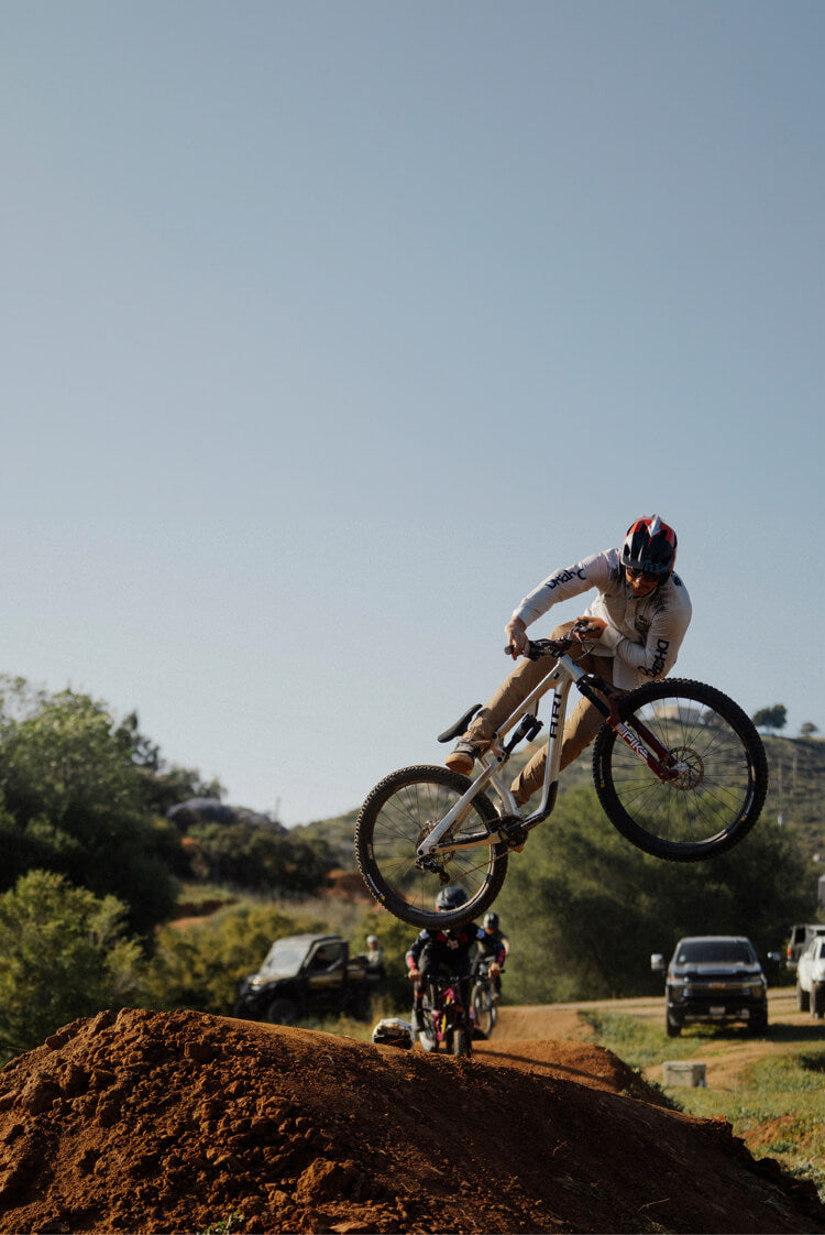 Kyle Strait performing a jump on a dirt track with vehicles and trees in the background