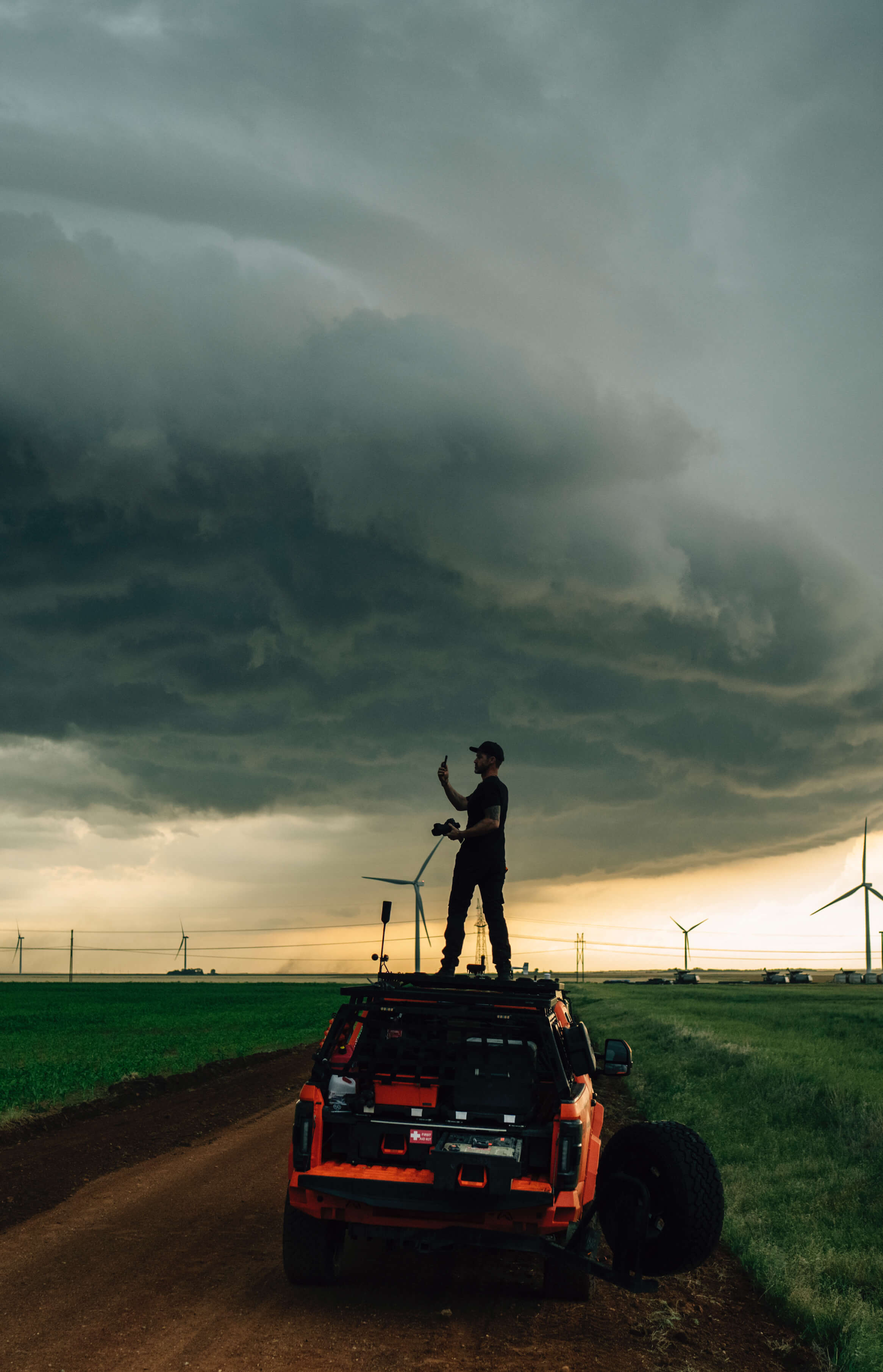 Ricky Forbes standing on top of his storm-chasing truck with a stormy sky and wind turbines in the background.