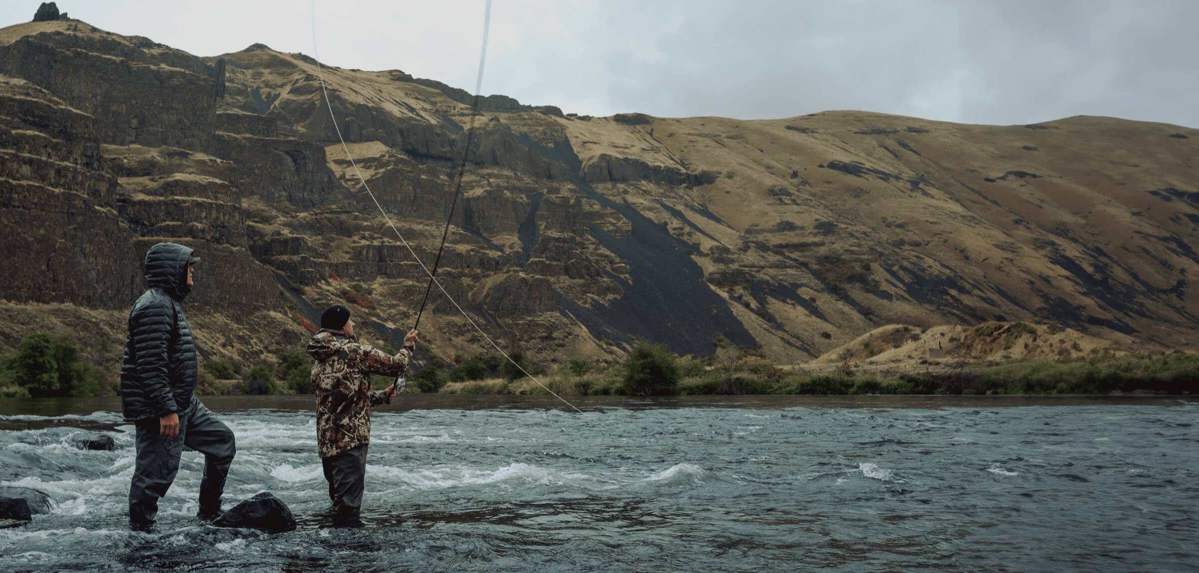 Two people fly fishing in a river with mountains in the background