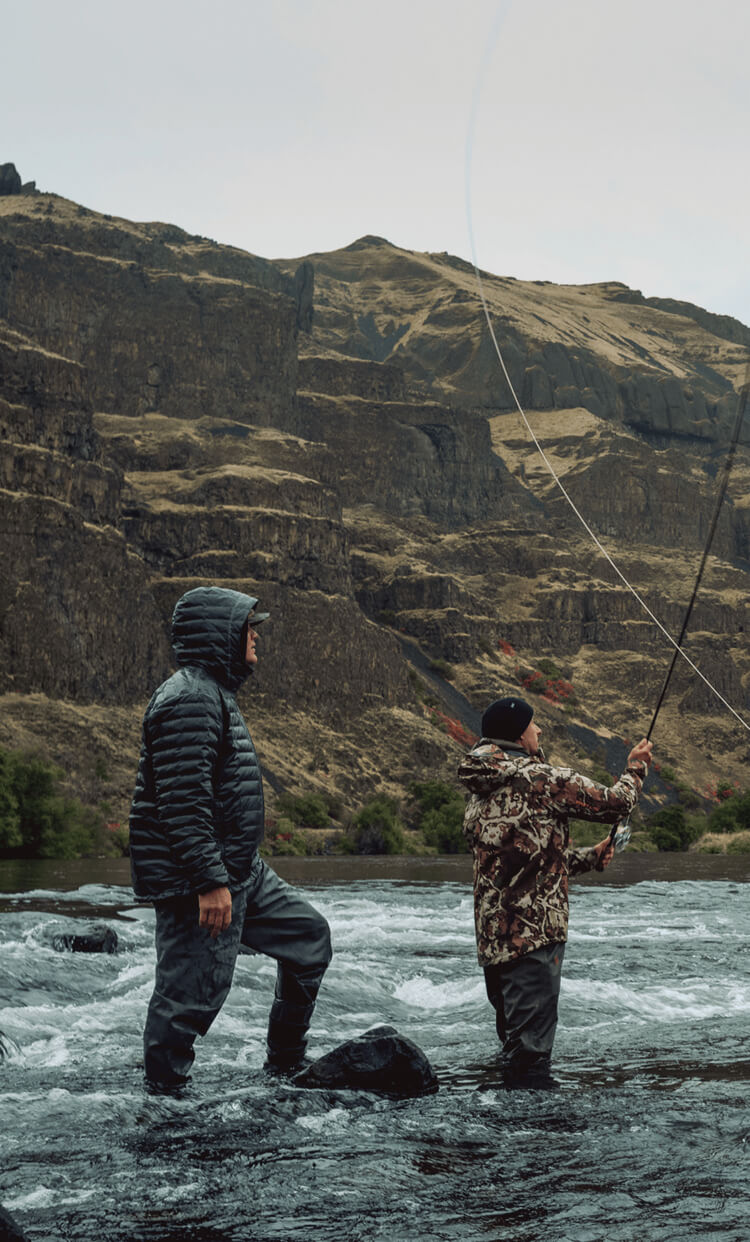Two people fishing in a river with mountains in the background