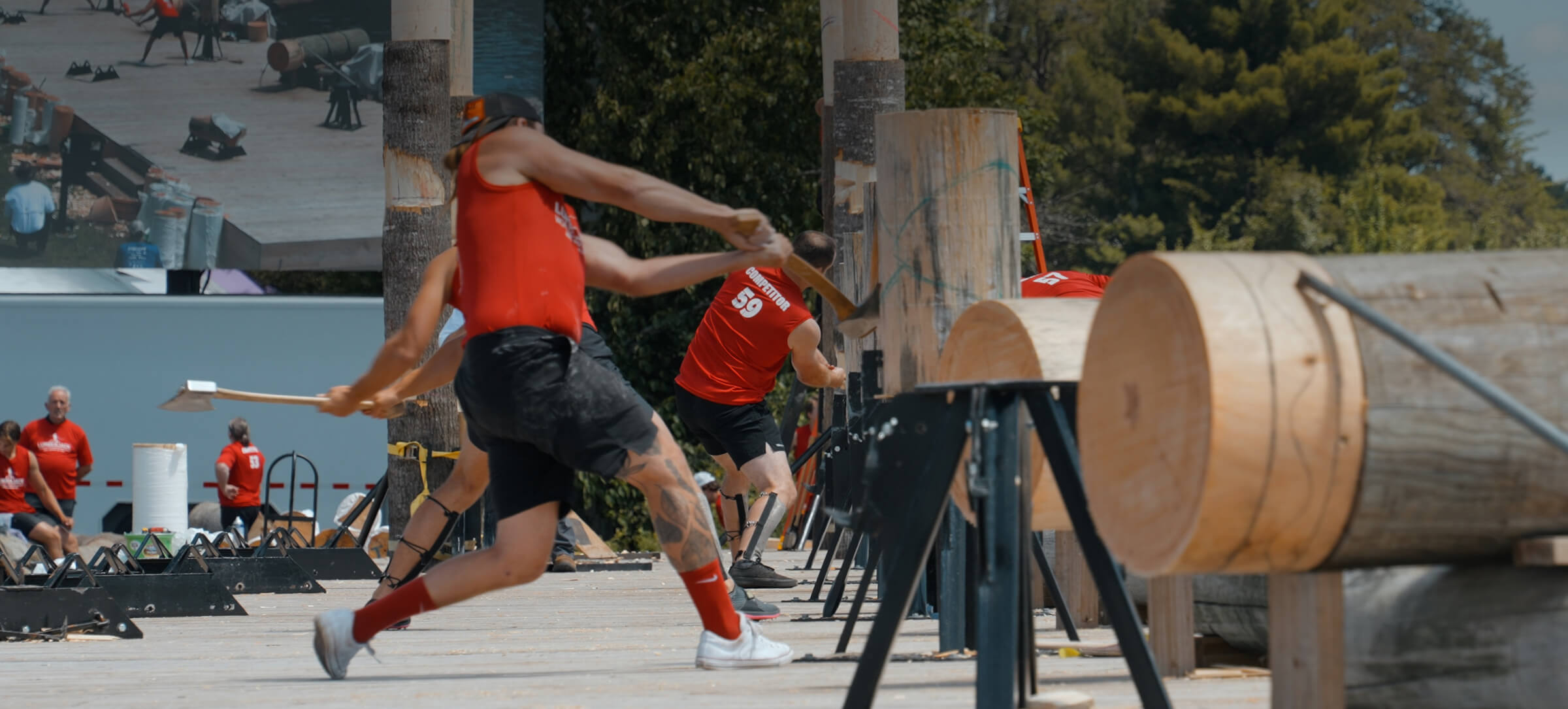 Two athletes in red athletic wear compete in a log-throwing event outdoors, with wooden targets and trees in the background.
