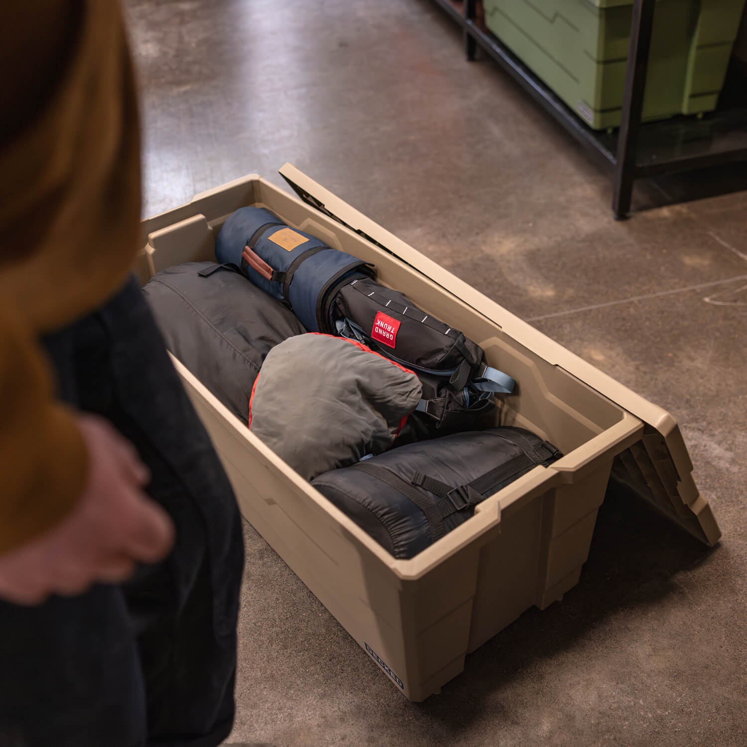 Person standing next to a large, weatherproof Payloader storage bin holding rolled-up sleeping bags on a concrete floor.