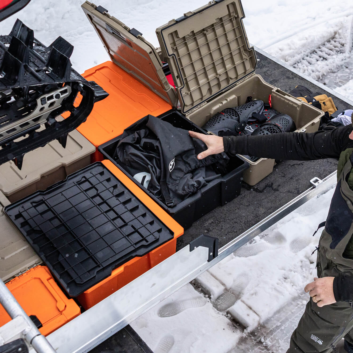 Person organizing snowmobiling gear in a Payloader case on top of a DECKED CargoGlide with snow-covered ground in the background