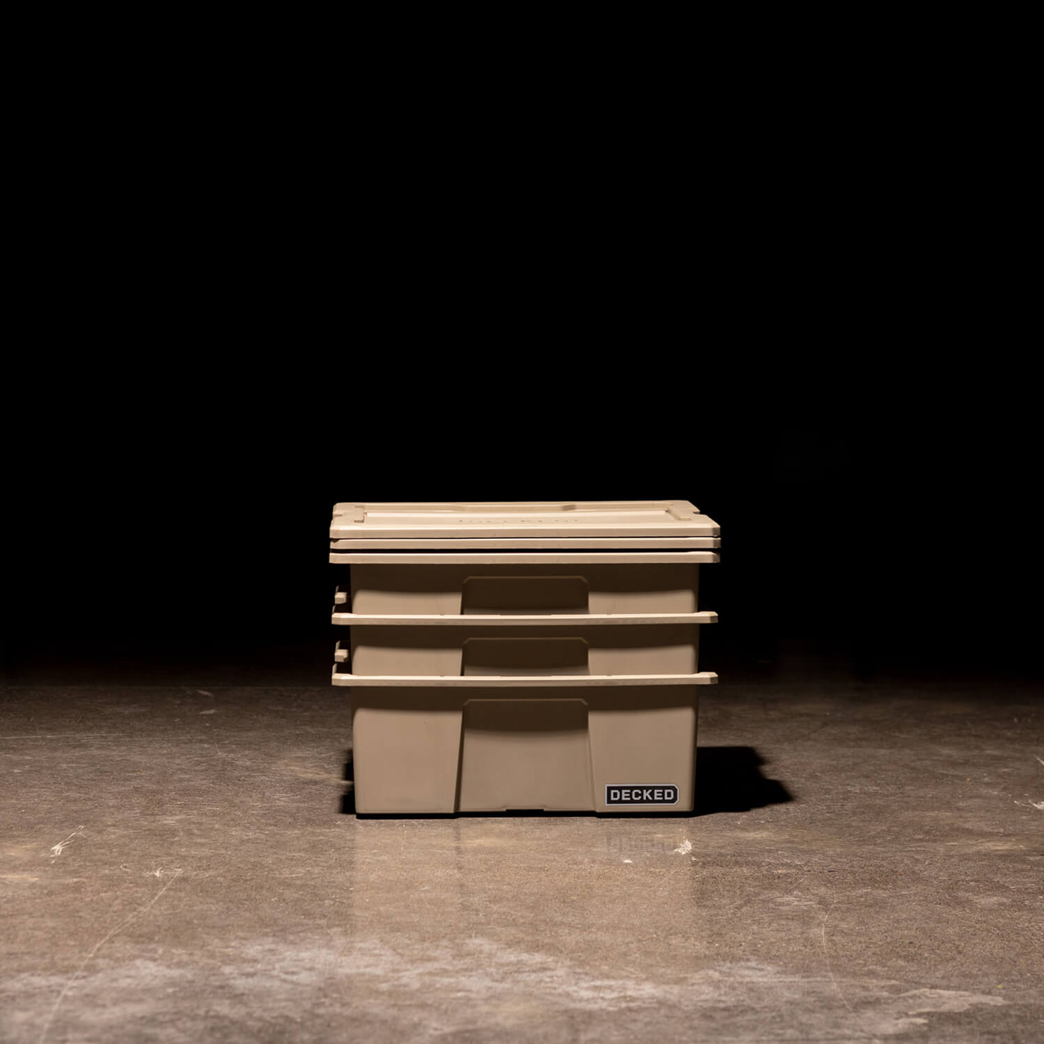 Three desert tan Payloader Bins nested together with the lids sitting on top in a garage with a dark, shadowy background.