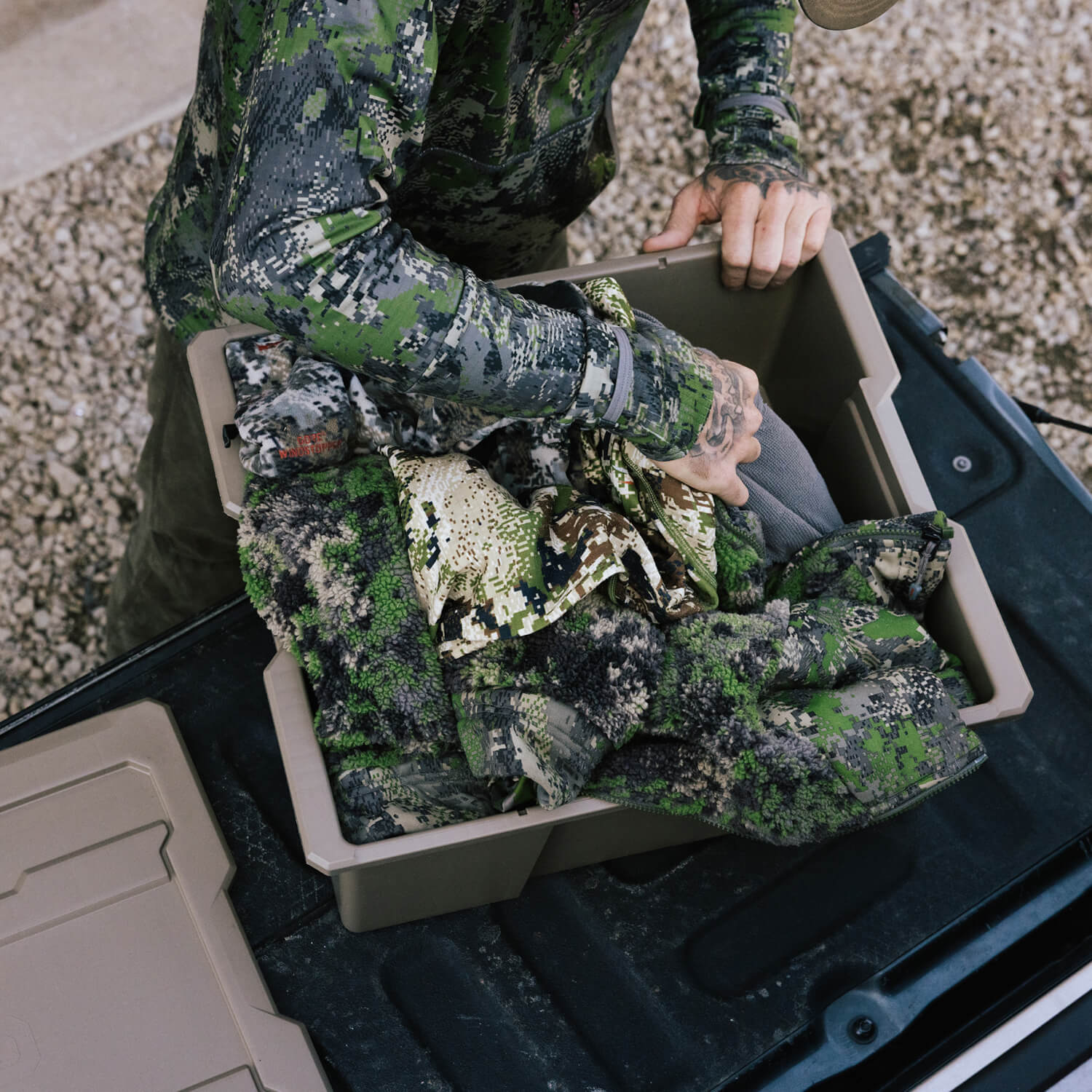 A man dressed in camo, packing his hunting outerwear into a desert tan Payloader Bin.