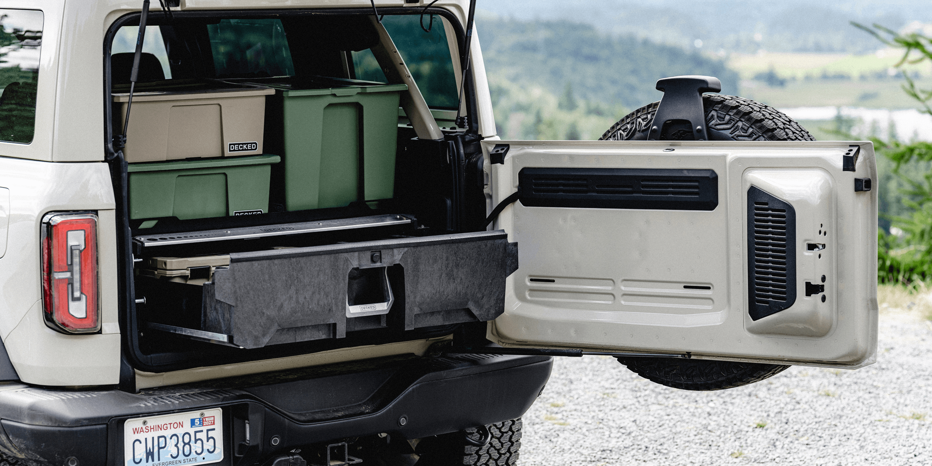 A white Ford Bronco SUV with its rear hatch and side door open, showing a built-in storage compartment and cooler system inside. The vehicle is parked outdoors on a gravel surface with a blurred natural background.