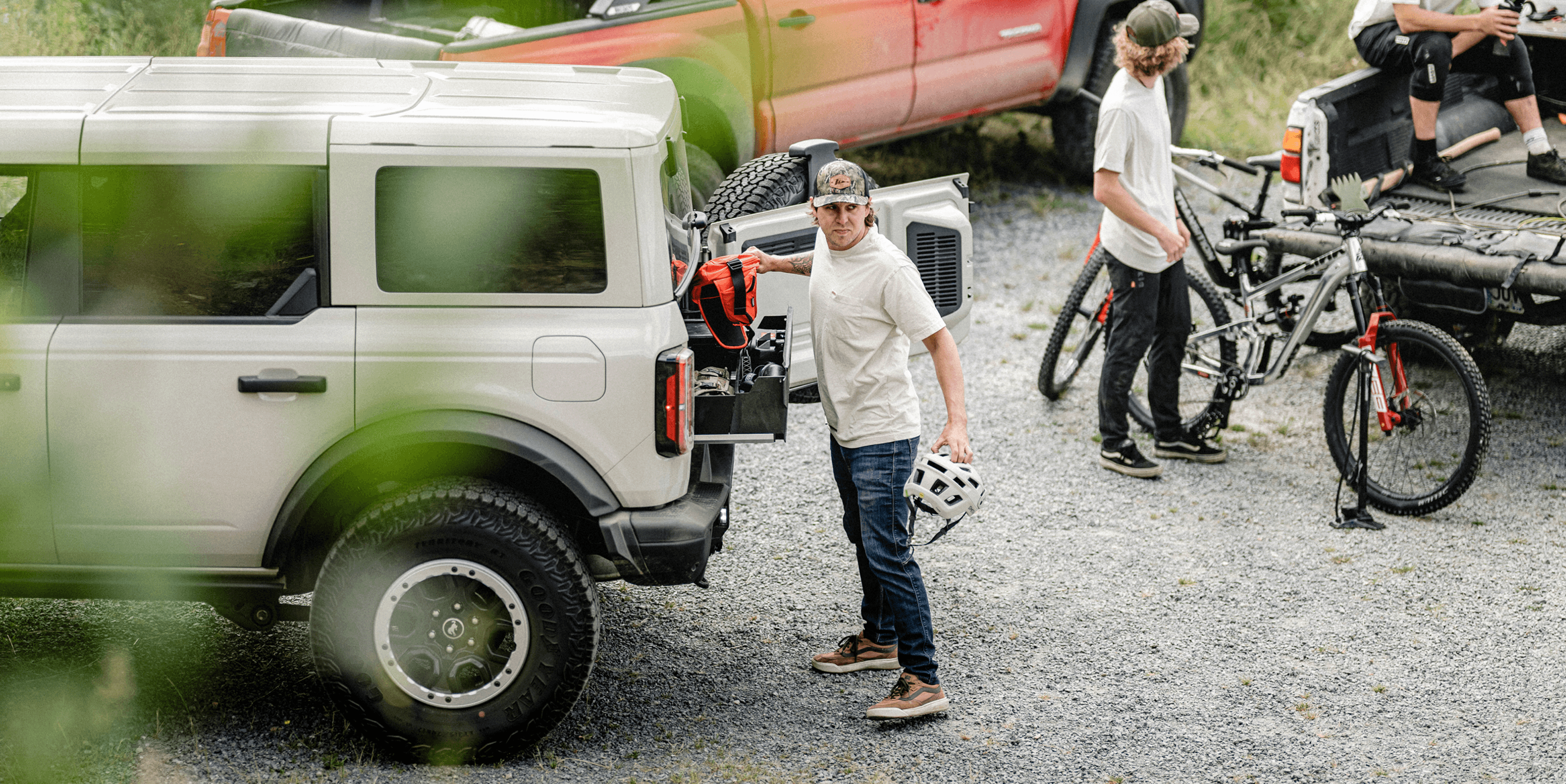 A man stands by the open rear hatch of a white SUV, holding a helmet, with bicycles and another person in a gravel outdoor setting.