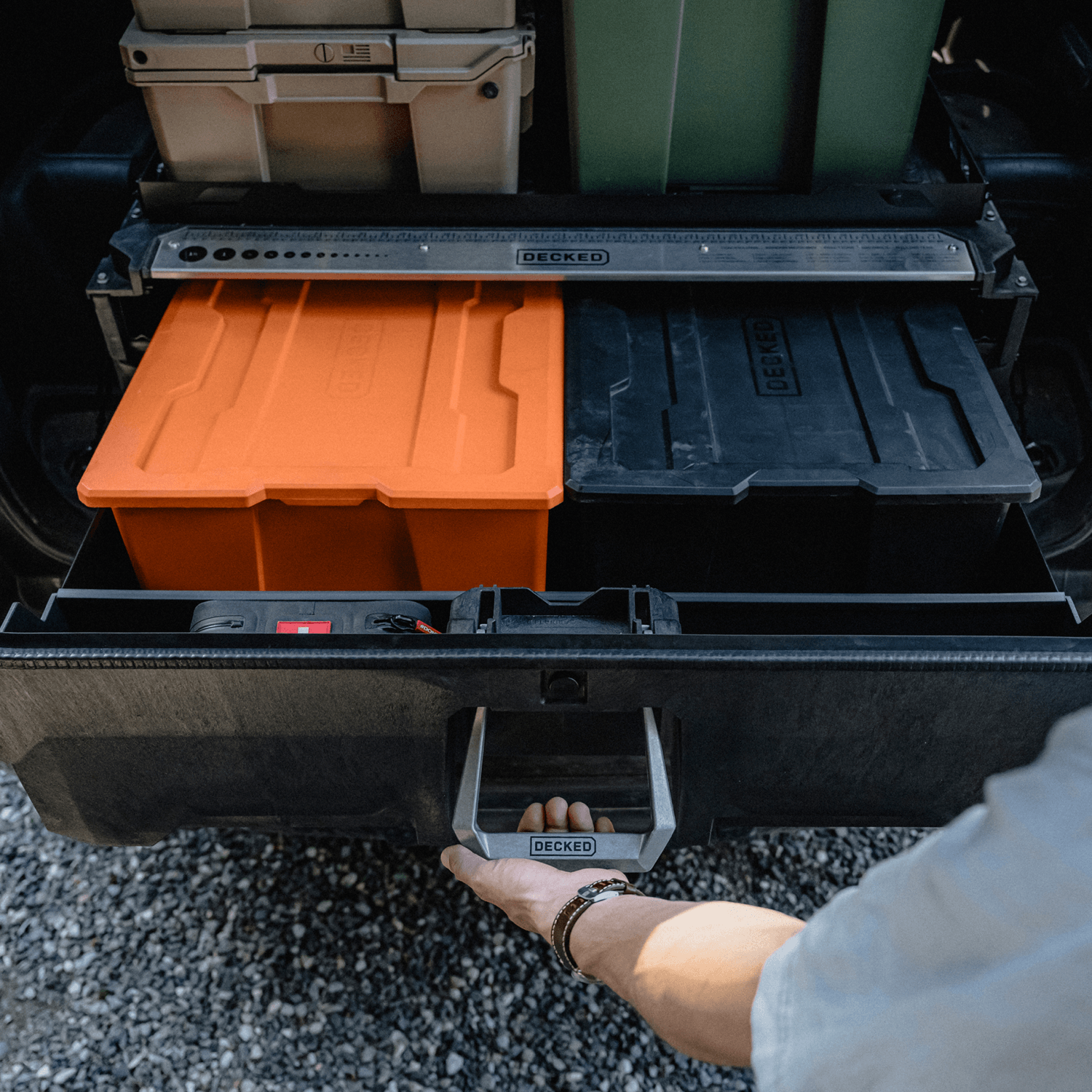 A person's hand is shown opening the drawer of a DECKED Drawer System which is open to reveal an orange storage bin and a black tray inside. Other DECKED bins are stacked in the background on a vehicle bed.