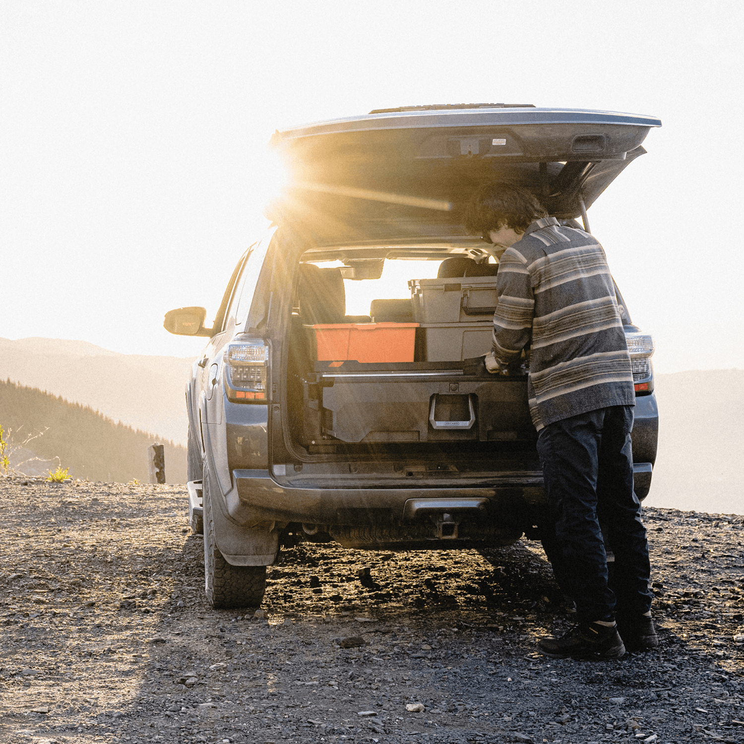 A man in a striped shirt stands by an open SUV tailgate on a dirt road at sunset, organizing gear inside the Drawer System in his trunk. Mountains are visible in the background.