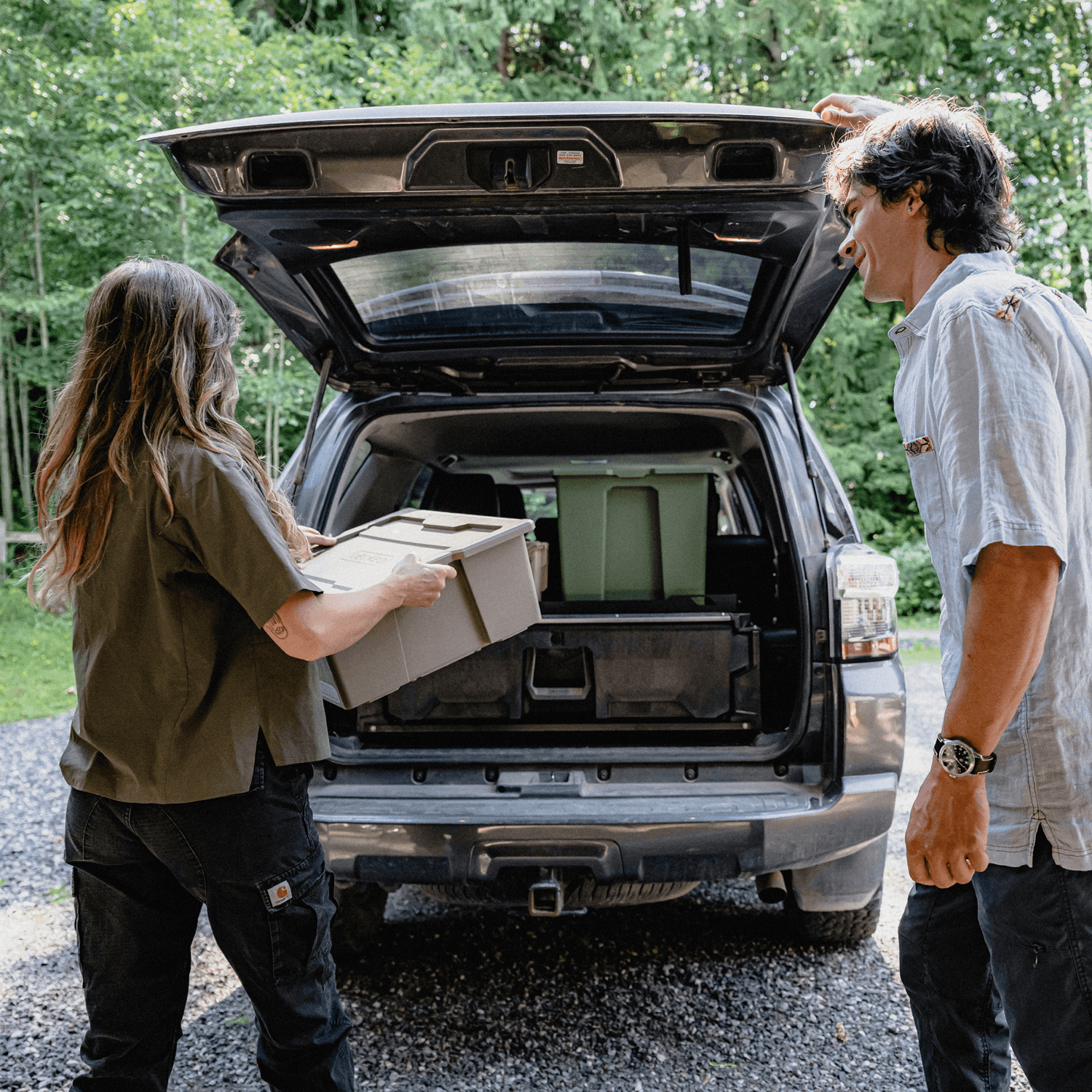Two people loading storage bins into the open trunk of a dark SUV with a Drawer System inside in a wooded outdoor setting.