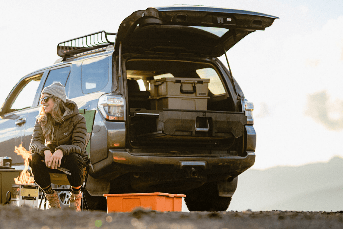 A woman sitting on a camp chair next to her 4-Runner Drawer System loading with camping gear.