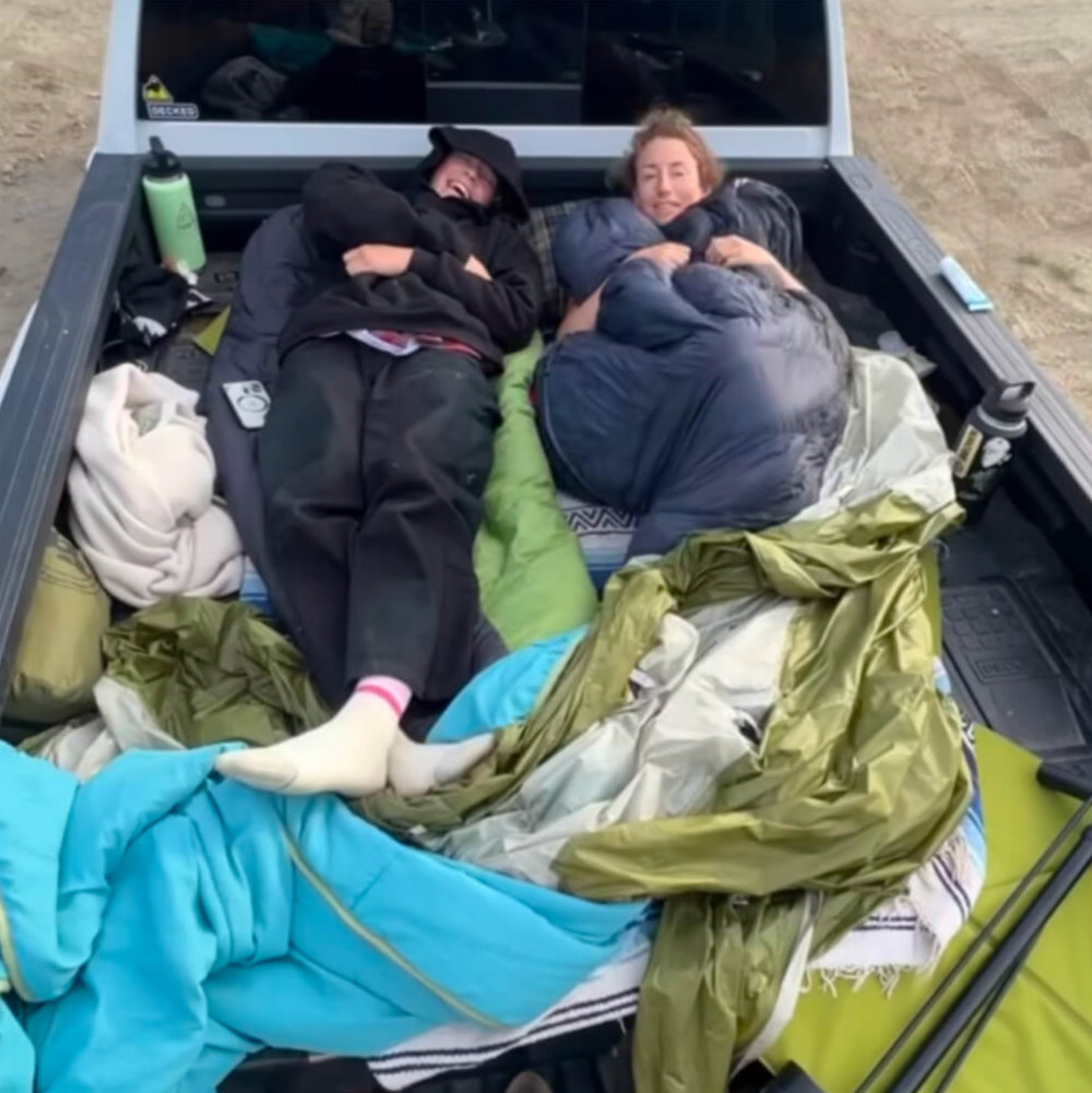 Casey and a friend lying on sleeping bags in the back of a truck with camping gear.