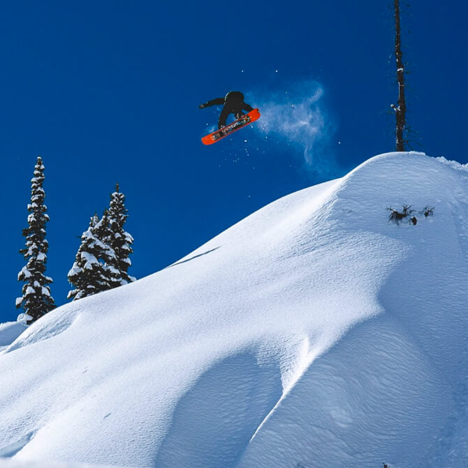 Curtis snowboarding off a cliff with a clear blue sky and snow-covered trees in the background