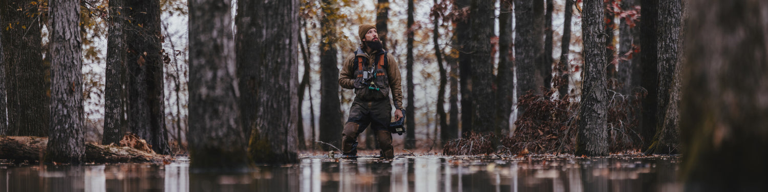Wade standing in the flooded timber in waders with a decoy in his hand and his duck calls around his neck