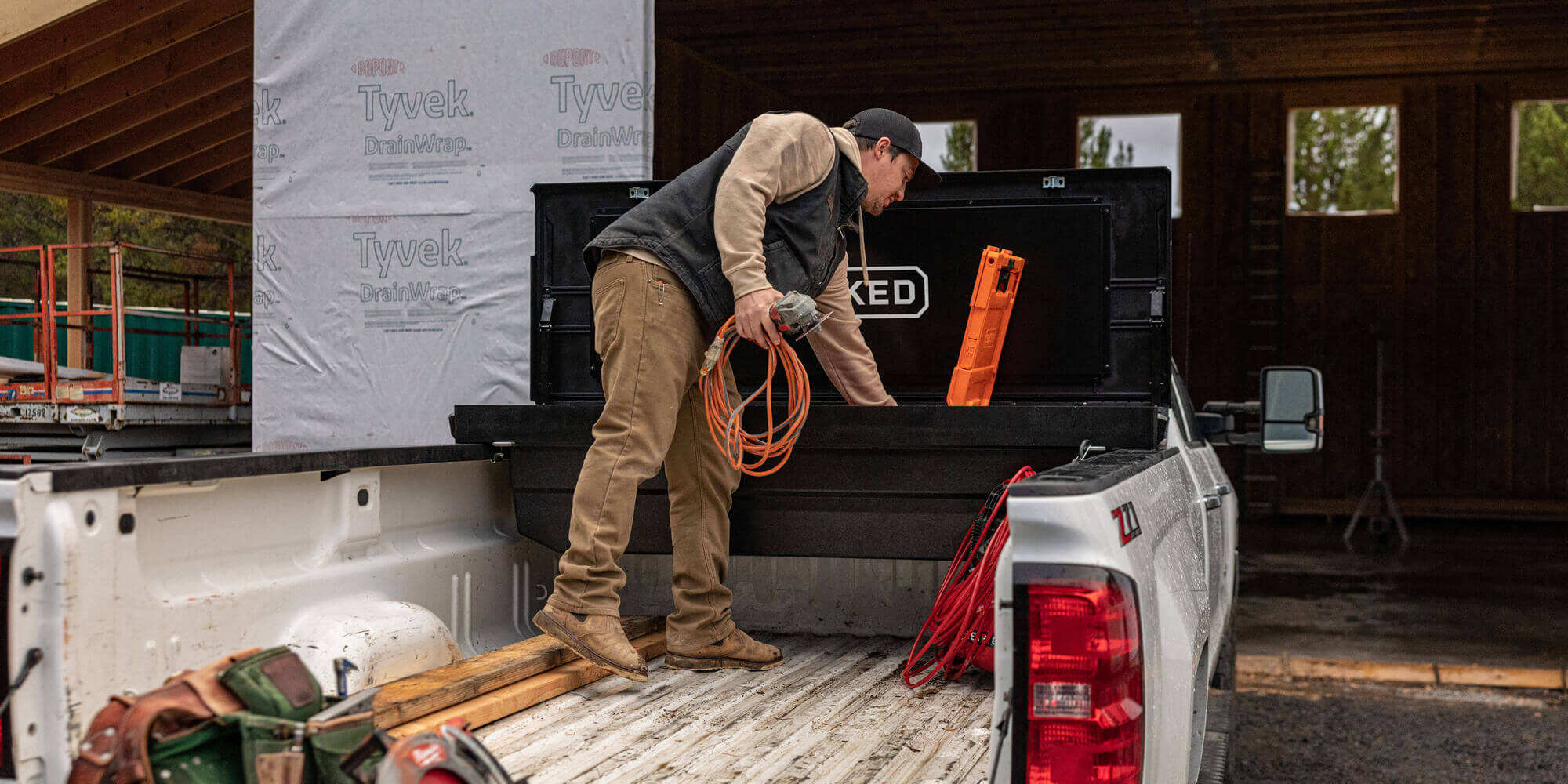 Person loading a black DECKED Tool Box in their truck bed with supplies, including a hose and a Halfrack D-co Case.
