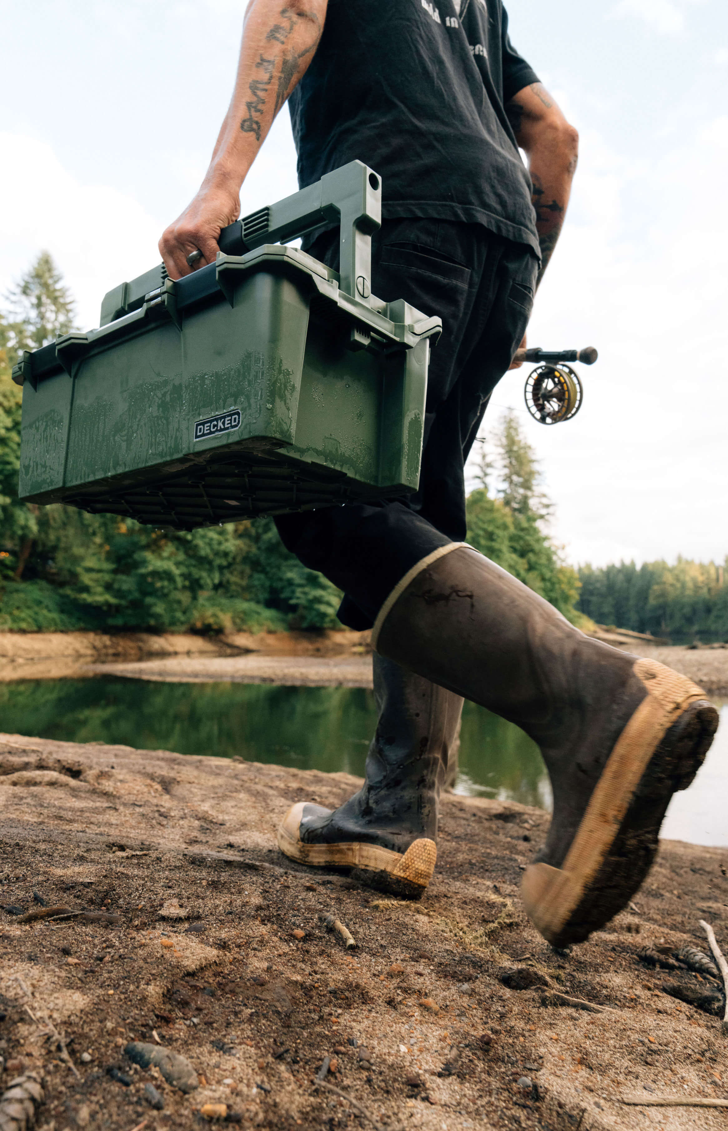 Person carrying a green D-co Case toolbox outdoors near a body of water
