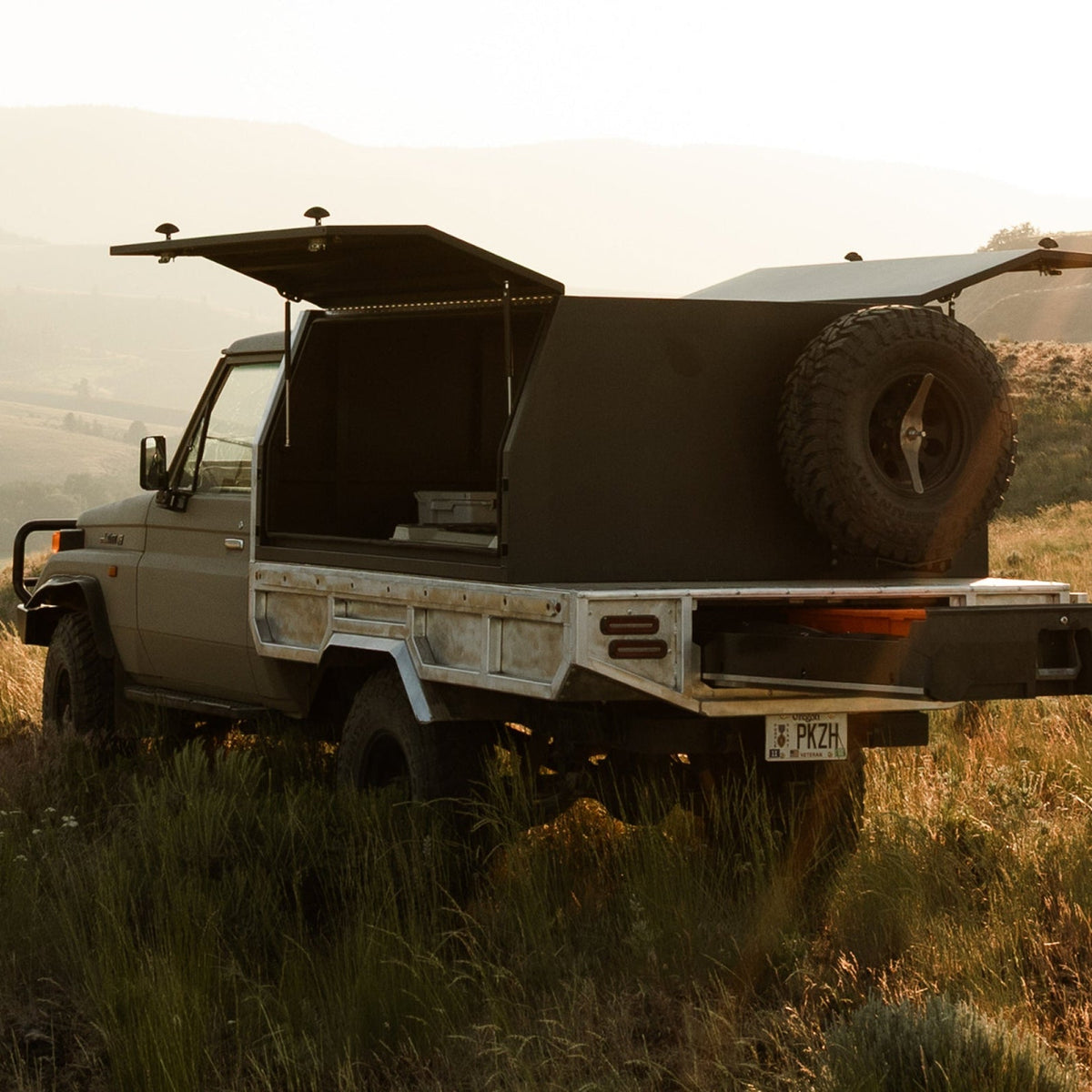 DECKED truck storage system mounted in the bed of a pickup truck parked in a grassy mountain landscape, with both side panels open and a spare tire mounted on the rear.