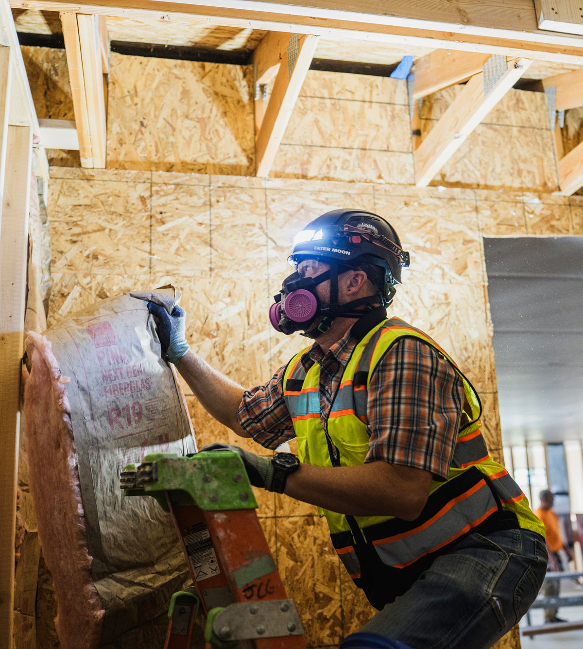 Construction worker wearing protective gear installs fiberglass insulation inside a wood-framed building.