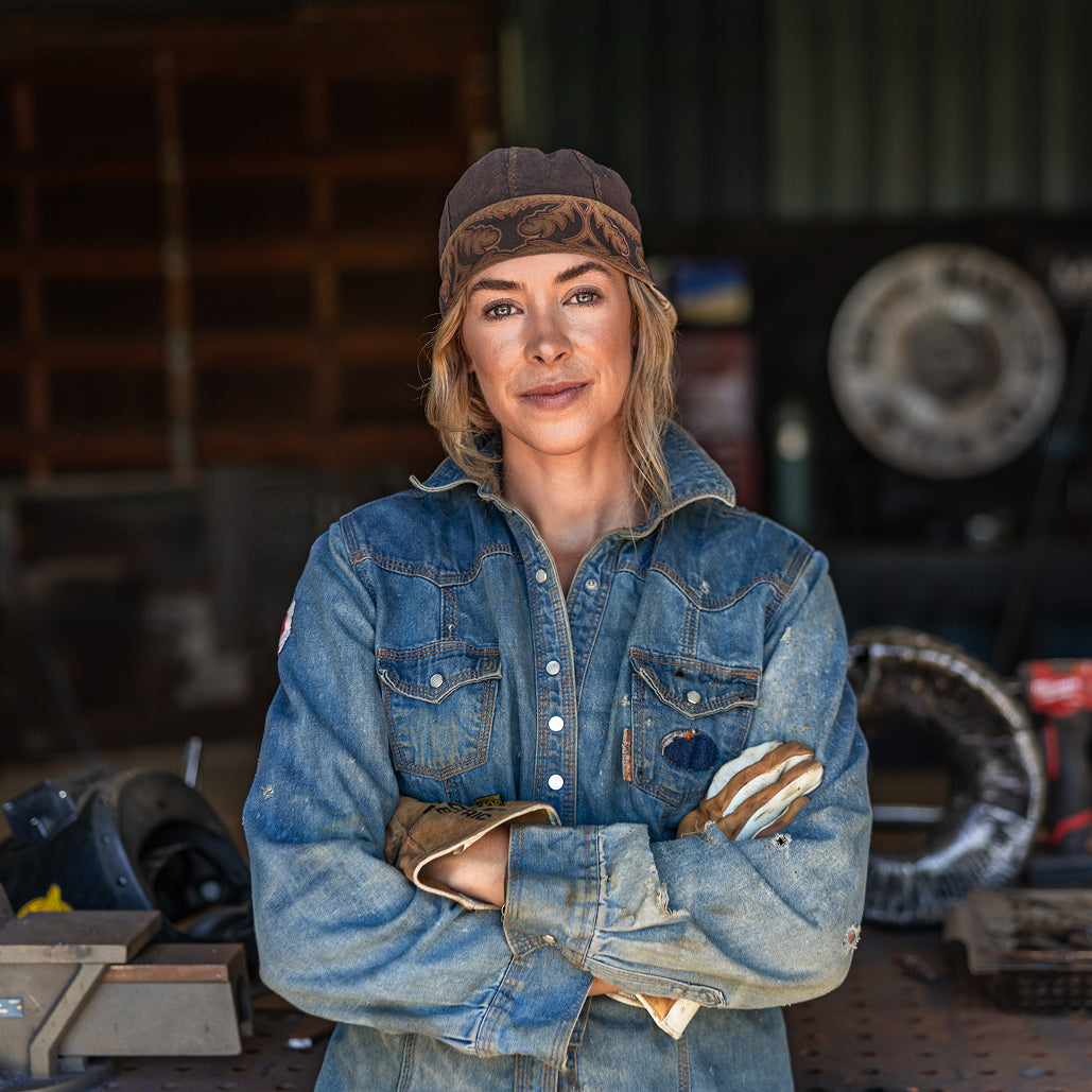 Female metalworker wearing a denim work shirt and leather gloves stands with her arms crossed in a fabrication shop.