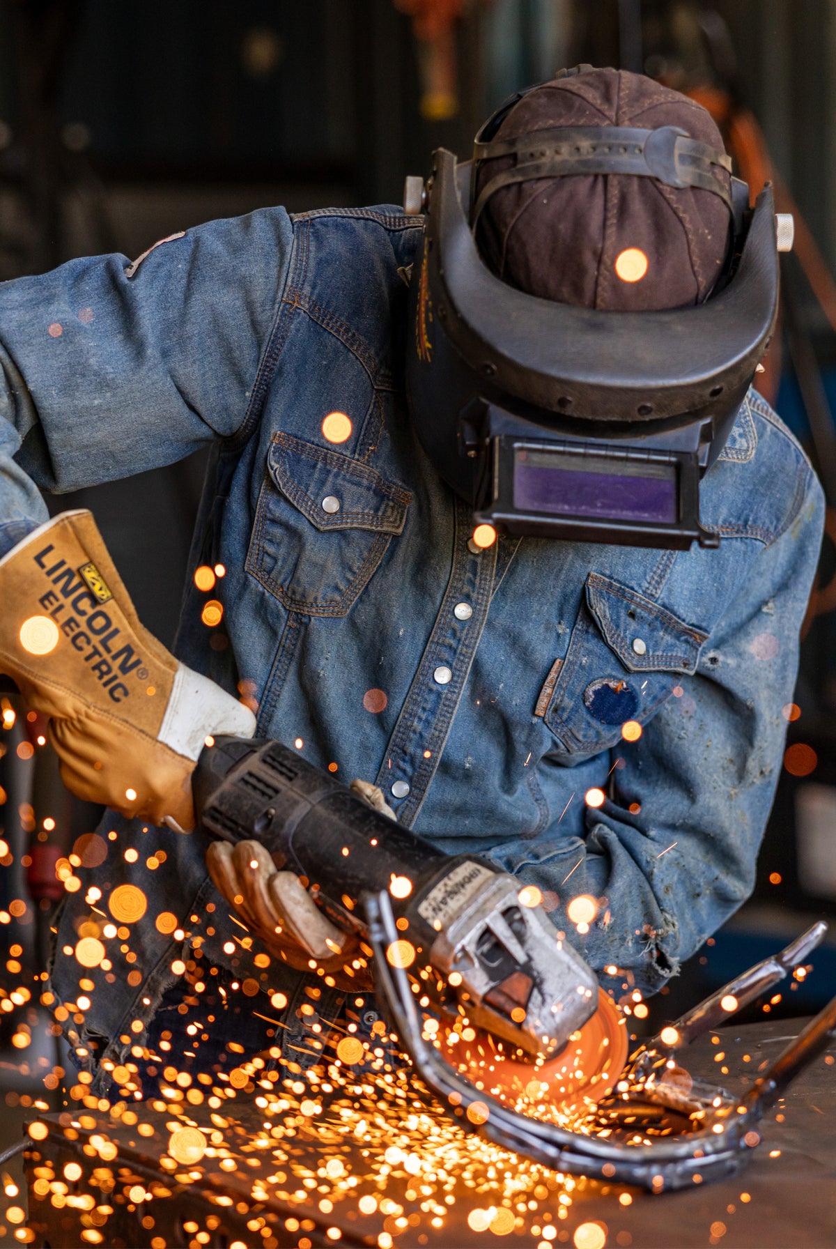 A welder in a blue denim work jacket and protective helmet sparks fly as they use a Lincoln Electric grinder, with bright orange sparks glowing in the foreground.