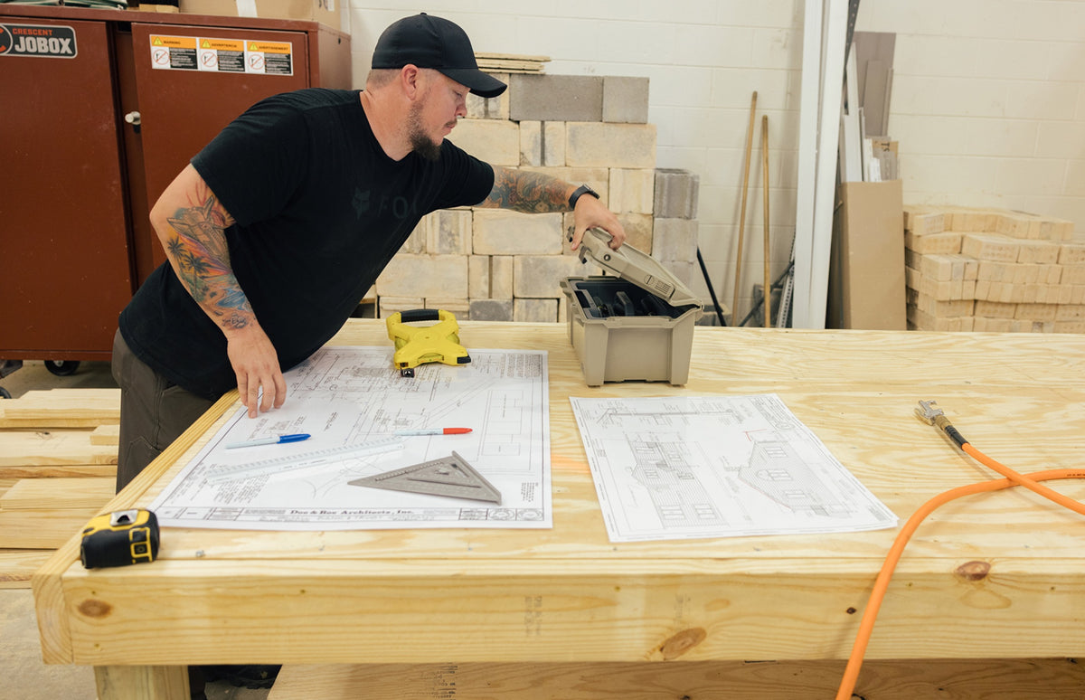 A builder opens a DECKED sixer case beside construction plans spread across a workbench.