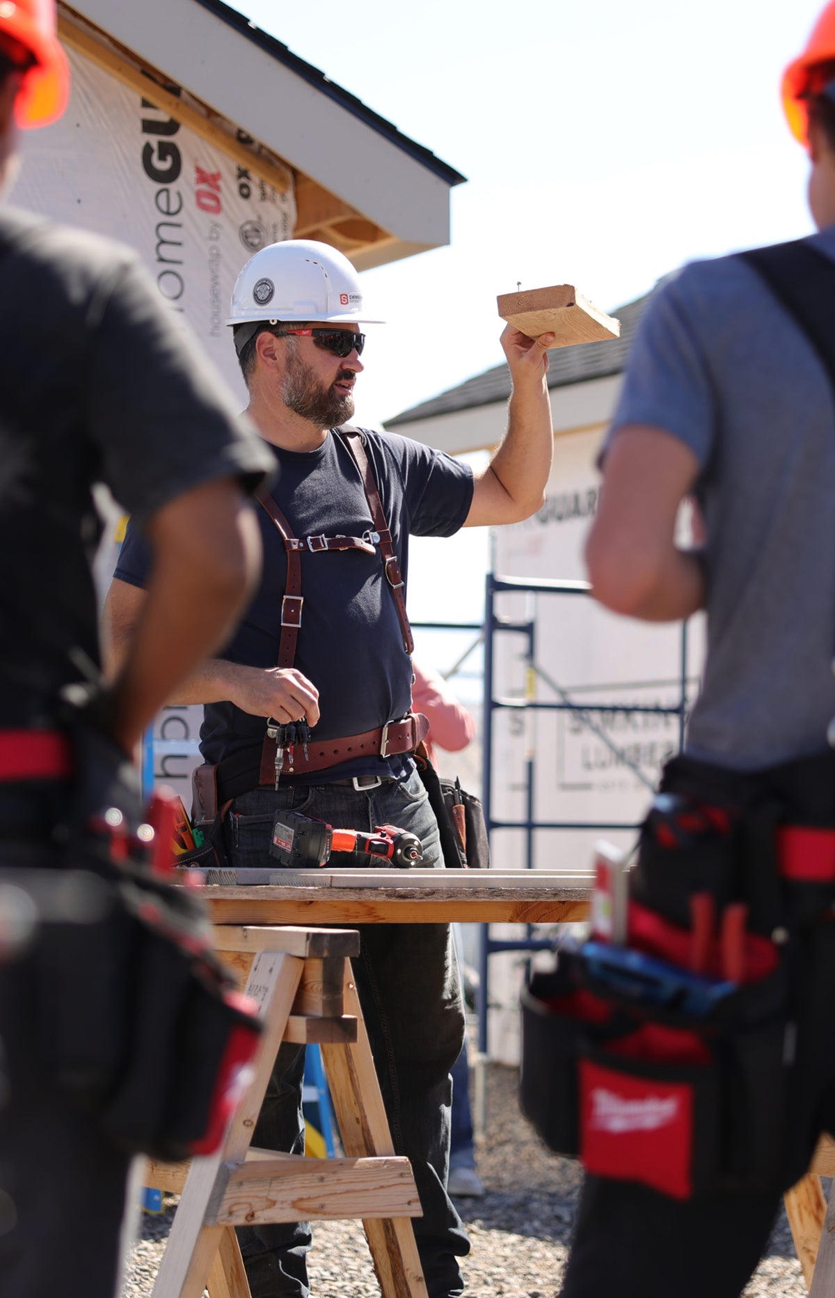 A carpenter holds up a piece of lumber while talking with two crew members at a construction site.