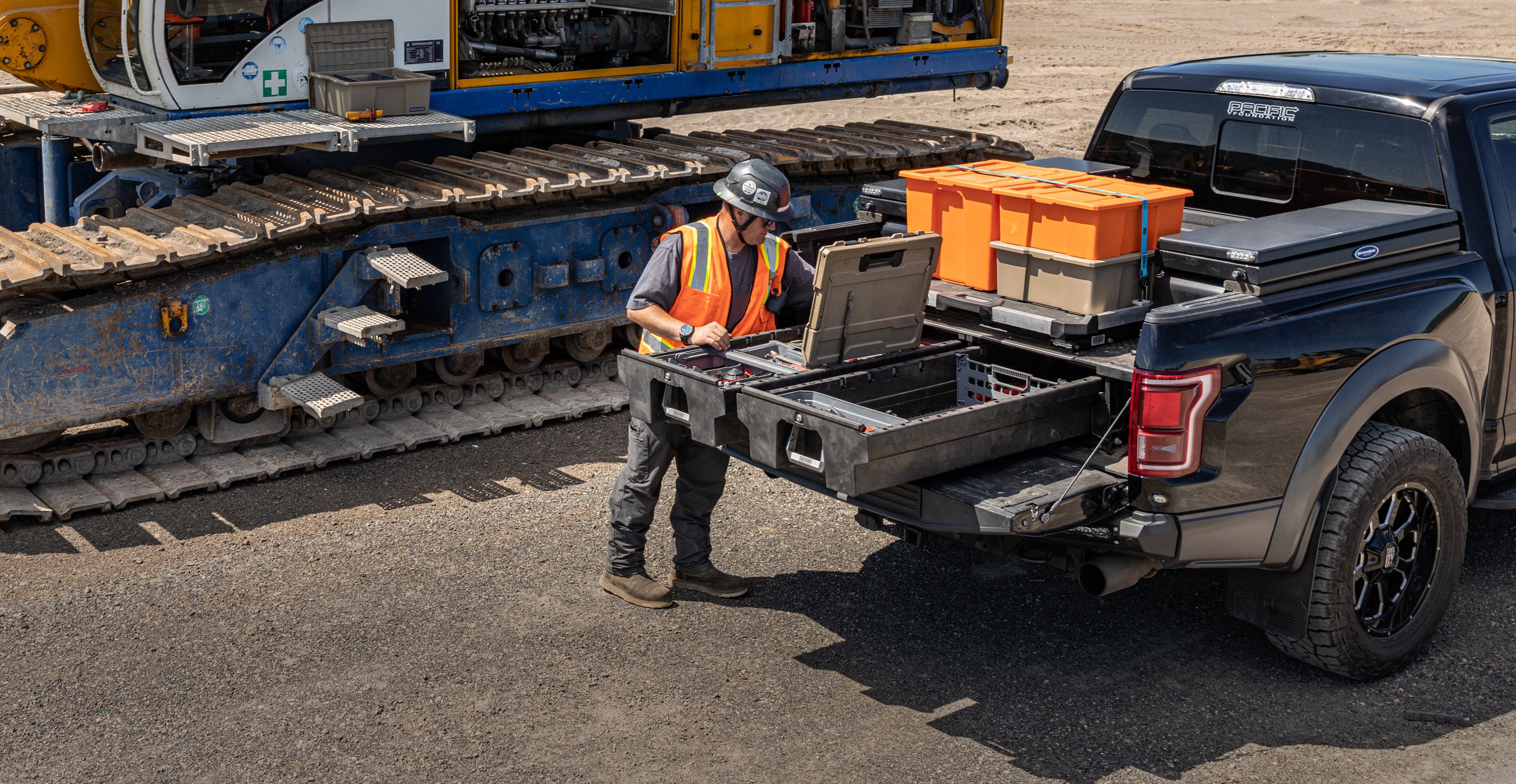 Construction worker accessing tools from a DECKED Drawer System and CargoGlide in a pickup truck bed beside heavy equipment at a construction site.
