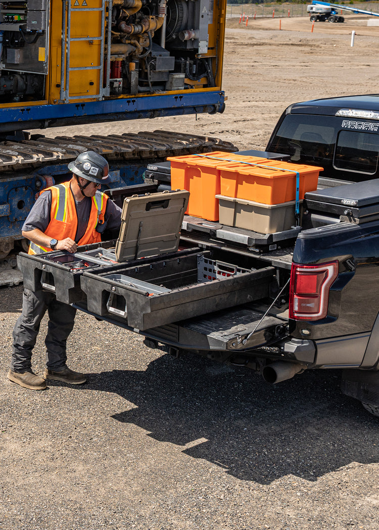 Construction worker accessing tools from a DECKED Drawer System and CargoGlide in a pickup truck bed beside heavy equipment at a construction site.