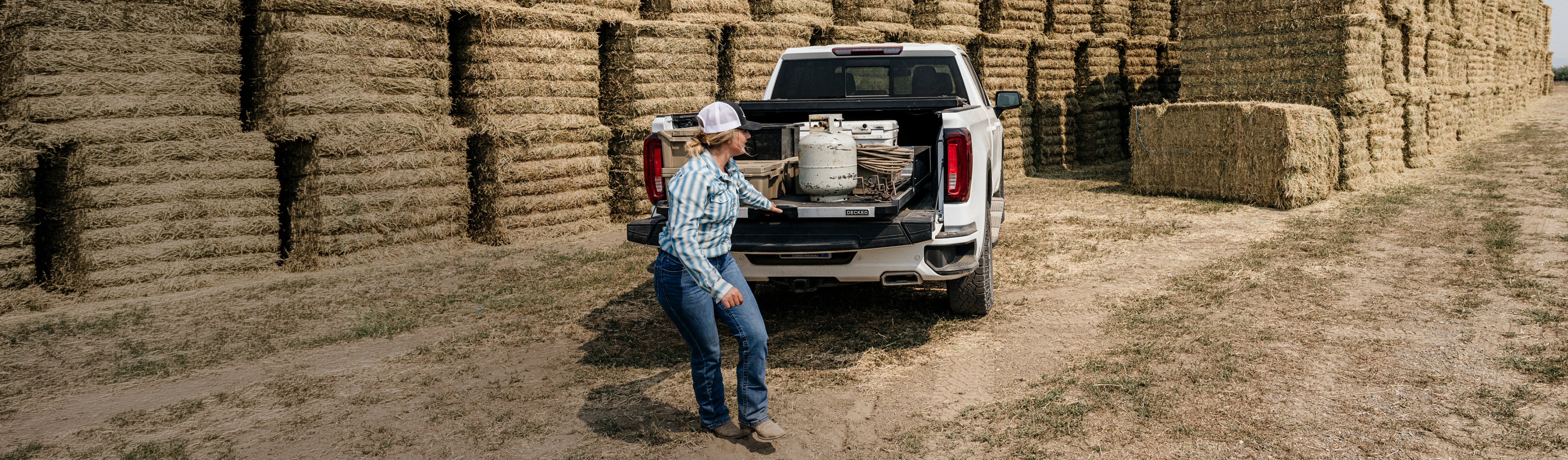 Rancher unloading equipment from a DECKED CargoGlide in a pickup truck bed beside stacked hay bales.