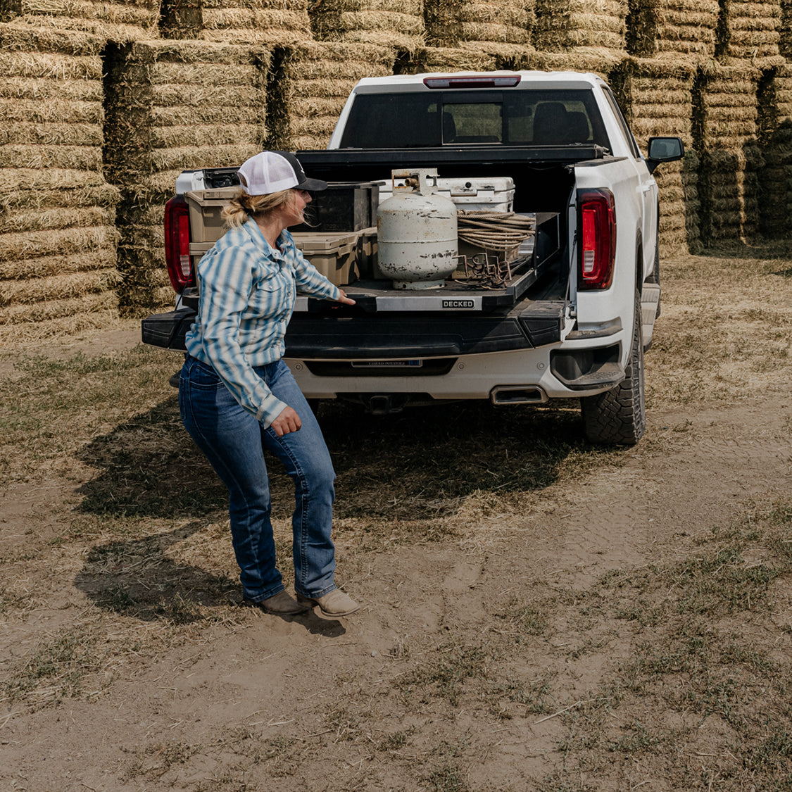Rancher unloading equipment from a DECKED CargoGlide in a pickup truck bed beside stacked hay bales.