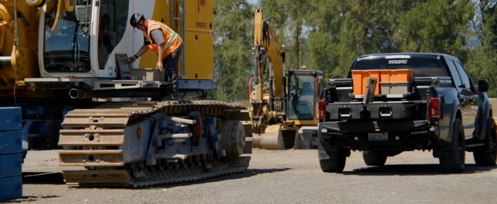 A black pickup truck with an Drawer System inside is shown from the rear in an outdoor construction site, alongside a large yellow tracked machine and a worker in an orange safety vest.
