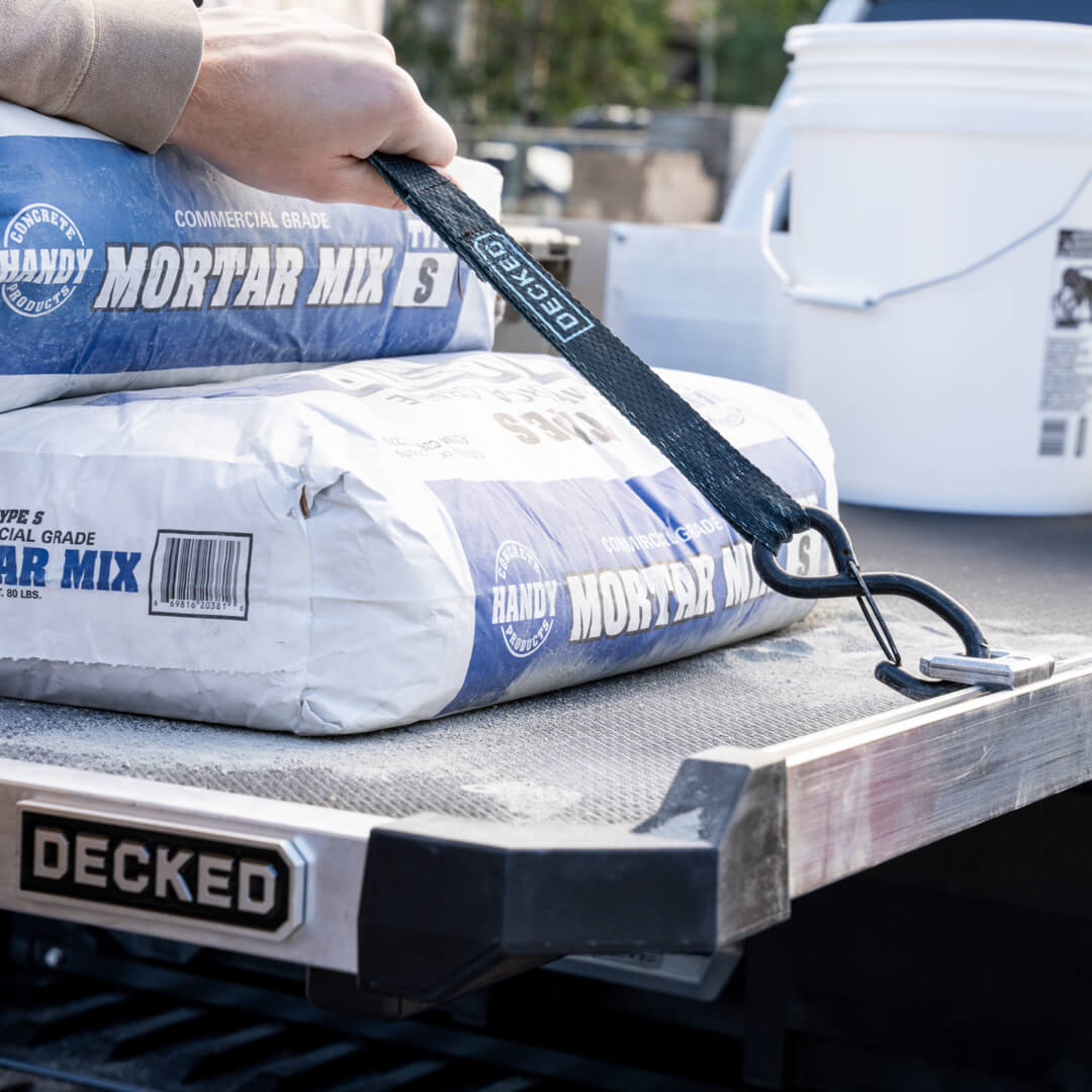 A DECKED cam strap attached to the D-rings for CargoGlide accessory holding down 80lb bags of mortar mix on top of a CargoGlide.